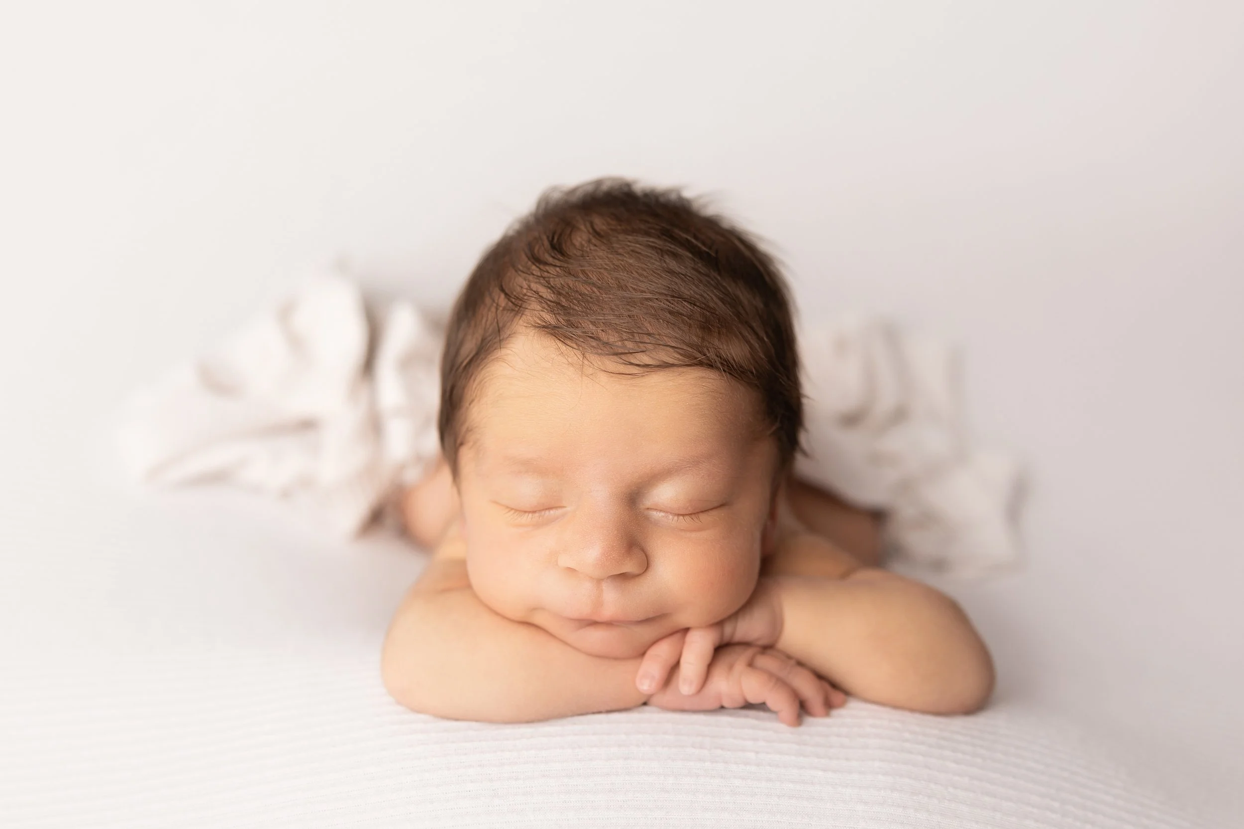 newborn baby boy resting chin on hands on white posing surface