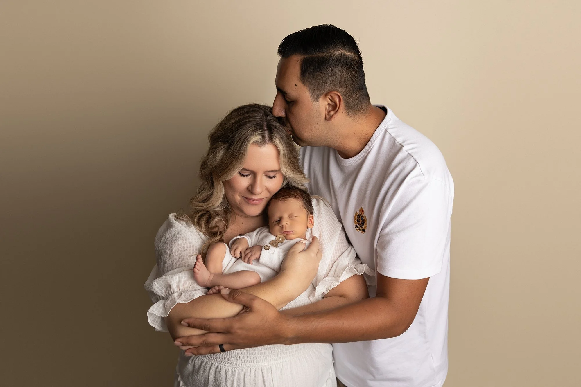 father gently kissing newborn baby while mother holds baby in studio portrait