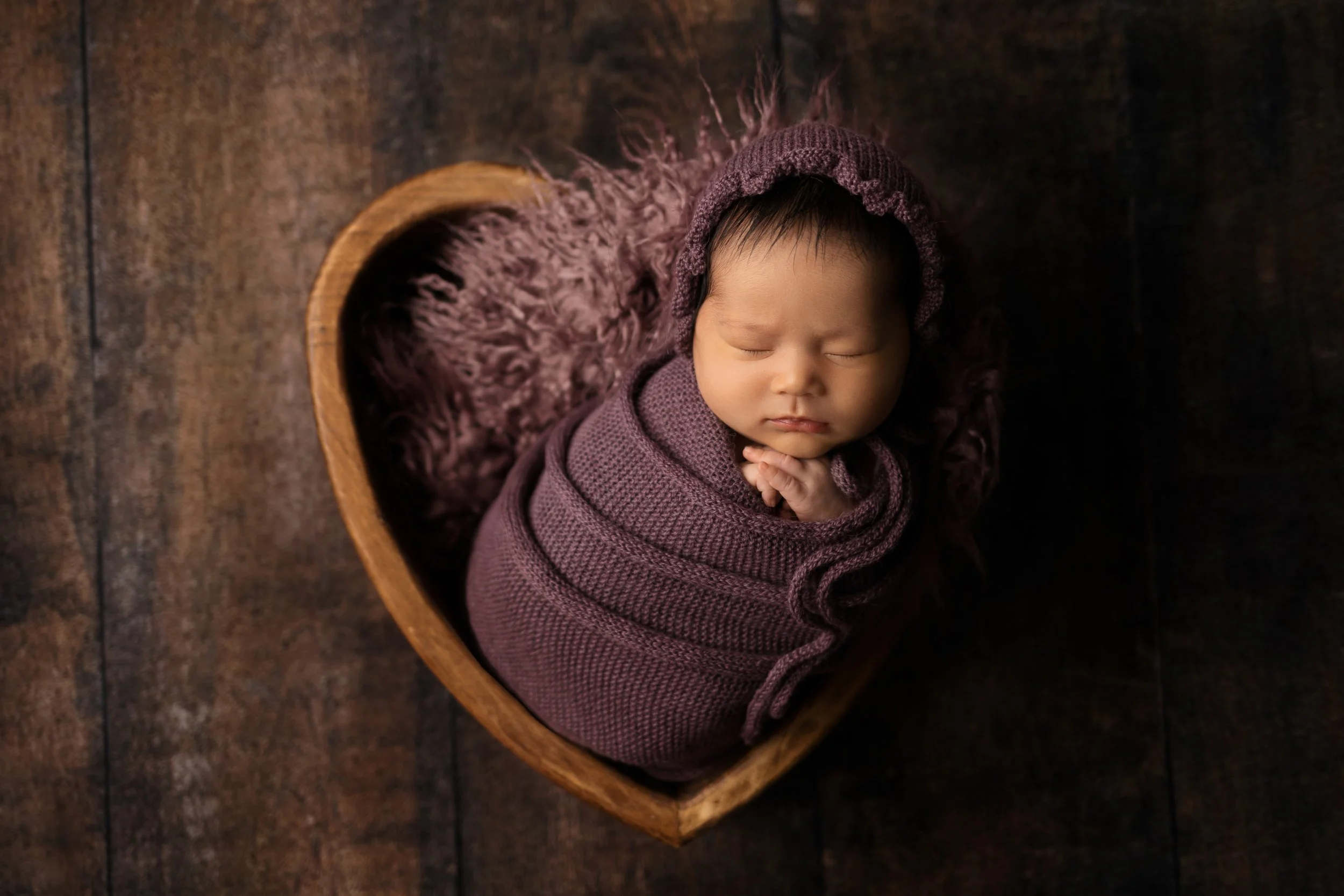 newborn baby girl wrapped in dark purple in heart shaped bowl posed in Austin Texas studio