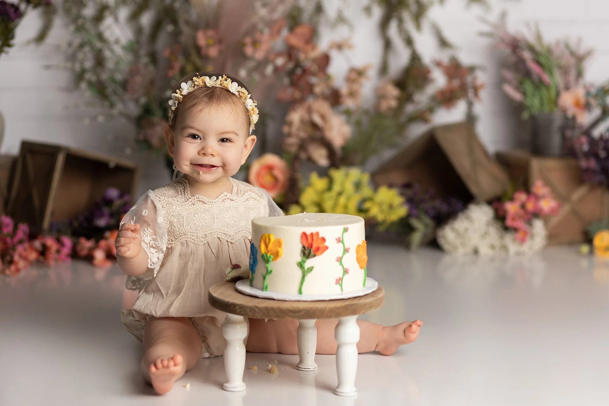 Smiling one year old girl sitting beside a floral birthday cake during a whimsical garden-themed cake smash session in Austin TX.