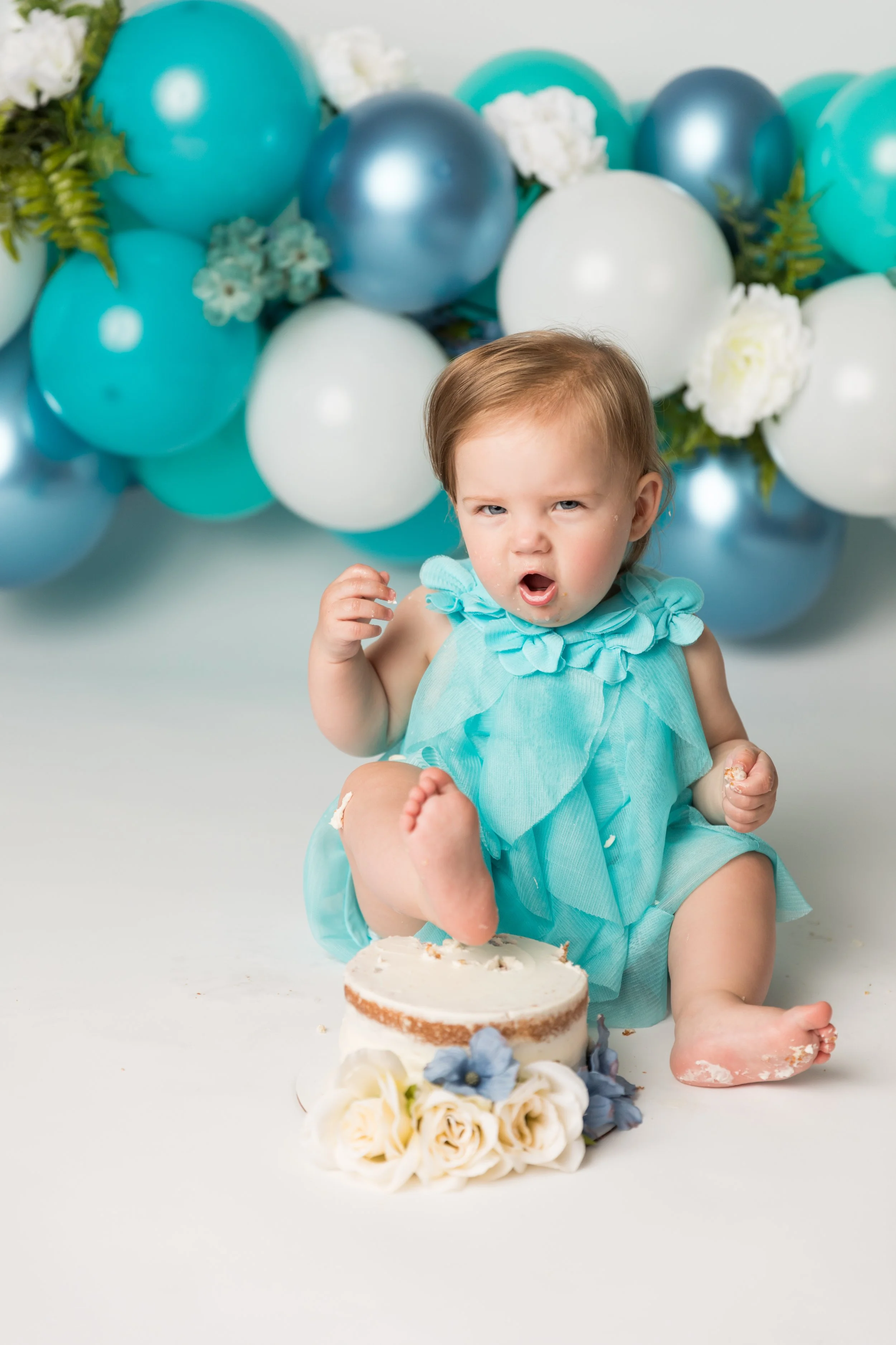 Baby girl enjoying her birthday cake during a blue, teal and white balloon-themed first birthday cake smash session.