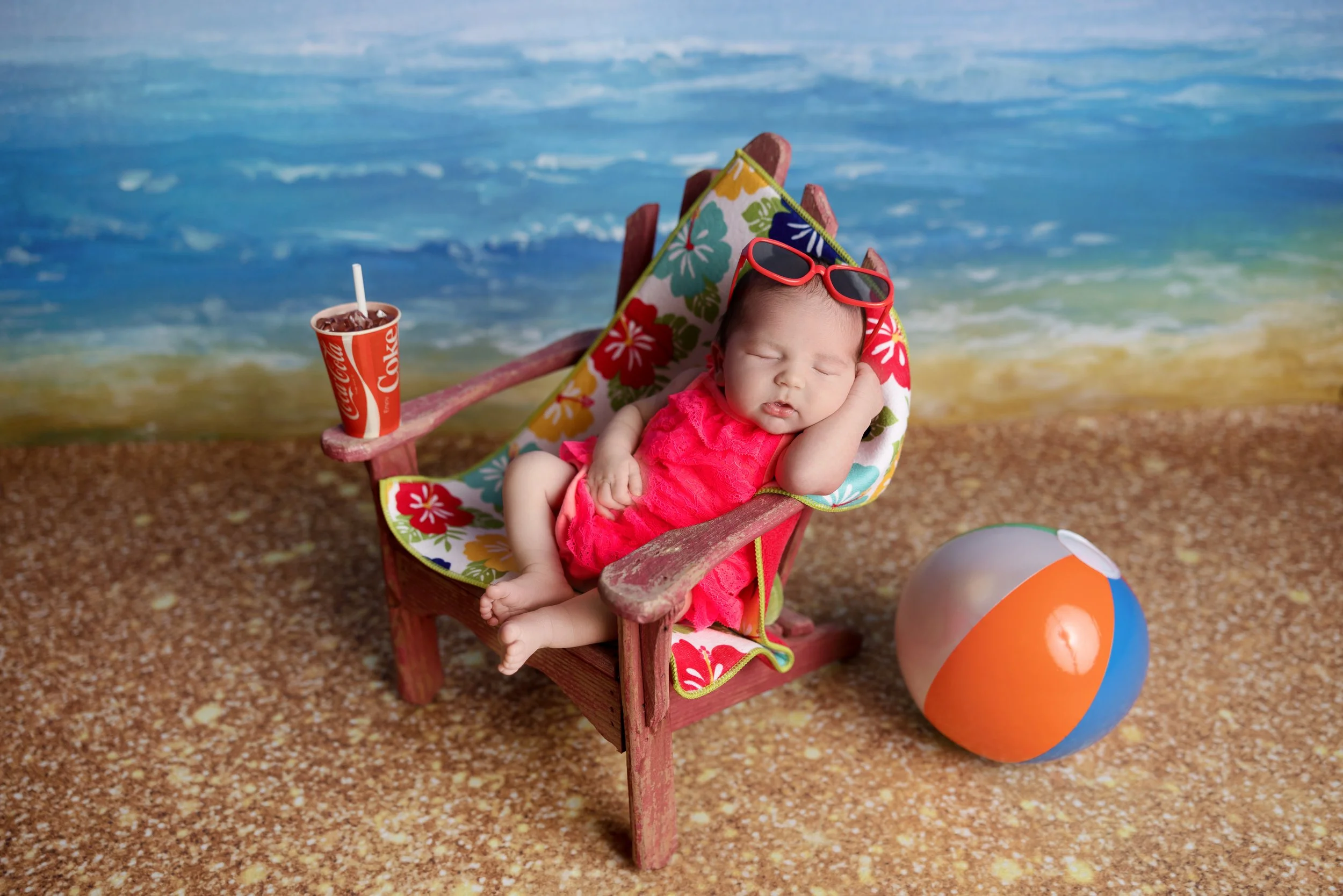 Newborn baby posed in a tiny beach chair with sunglasses, beach ball, and ocean backdrop for a summer-themed newborn photo session
