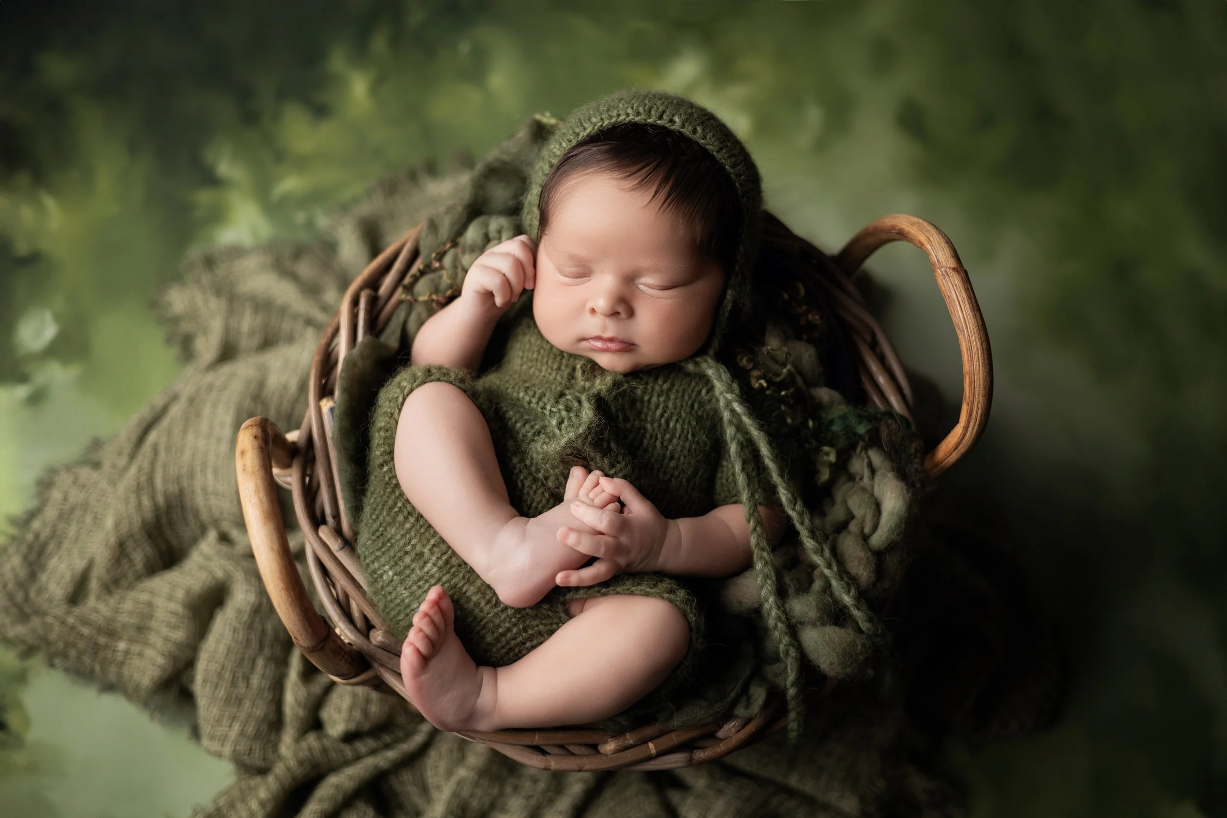 newborn boy in green knit romper and bonnet posed holding foot in basket with green background in Austin Texas studio