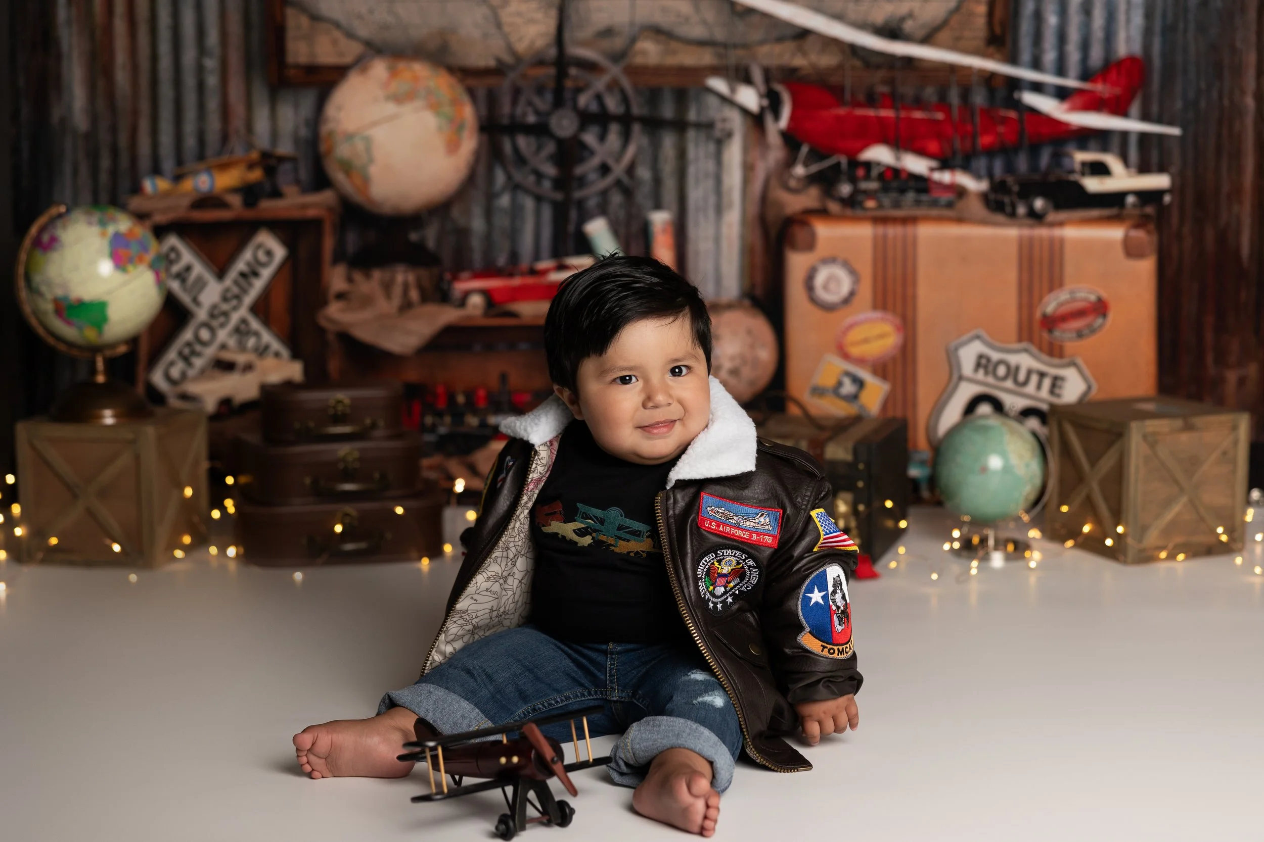 One year old boy in a vintage aviator jacket sitting with airplane props during a travel-themed cake smash session in Austin Texas.