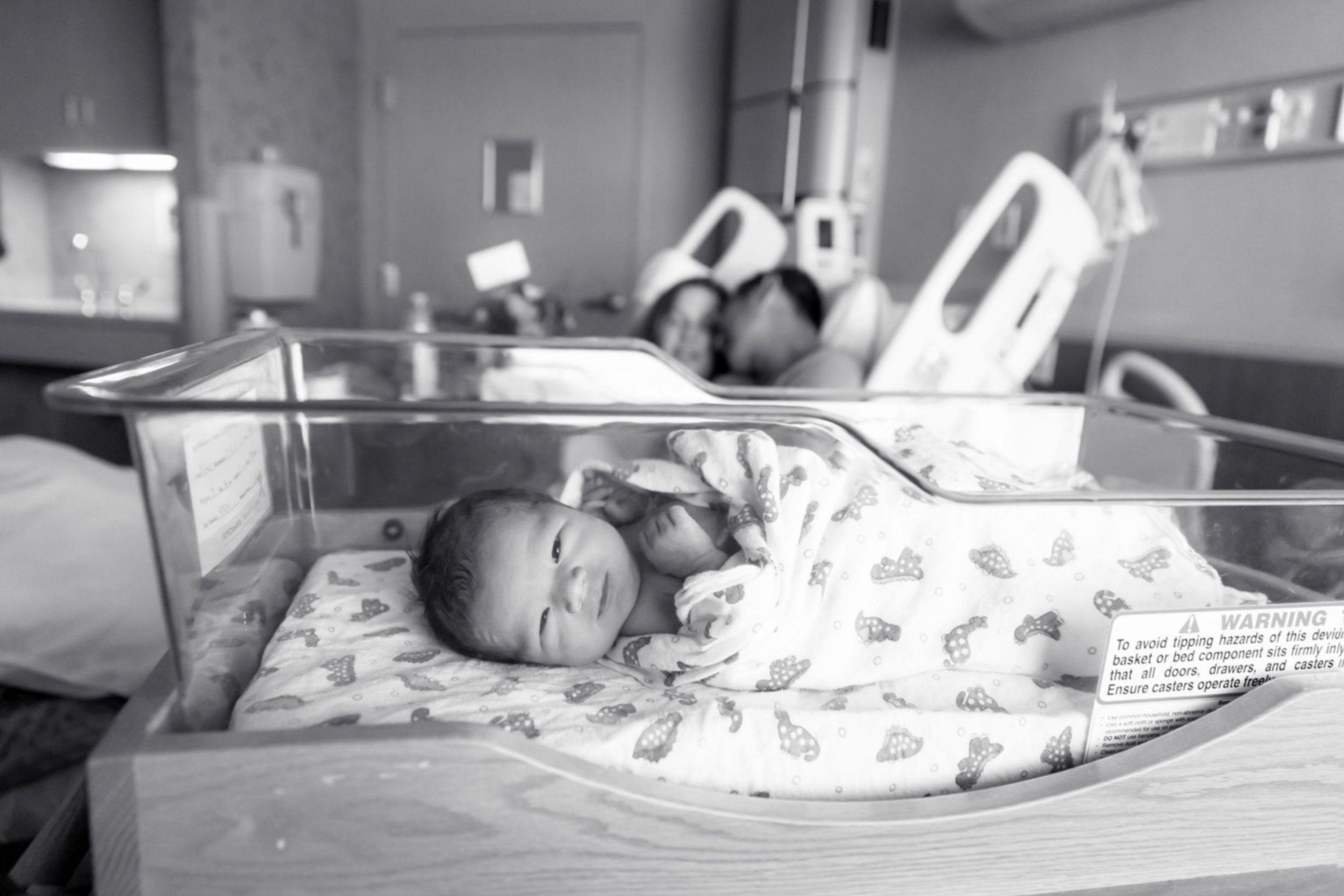 Newborn baby in hospital bassinet with parents snuggling in the background.
