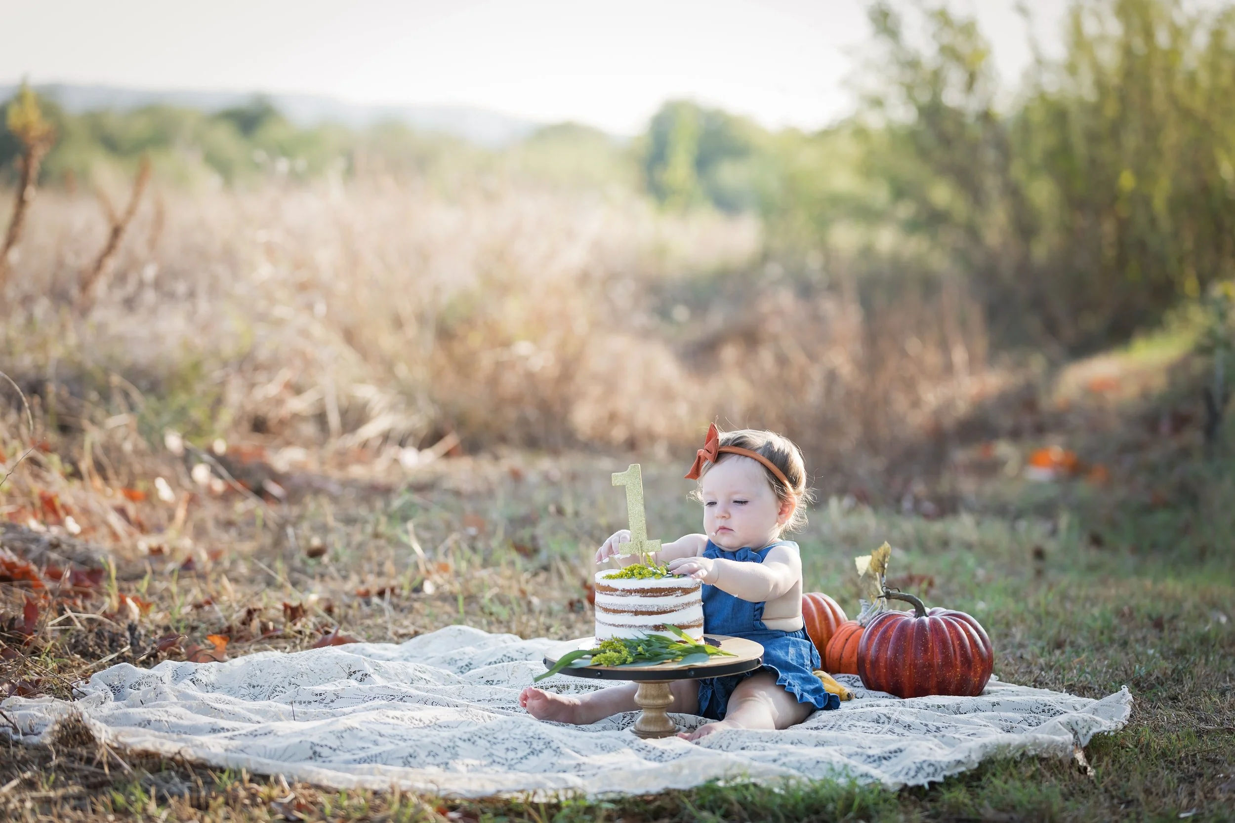 One-year-old girl sitting on a blanket with a simple first birthday cake and pumpkins during an outdoor fall cake smash session at Commons Ford Ranch in Austin, Texas.