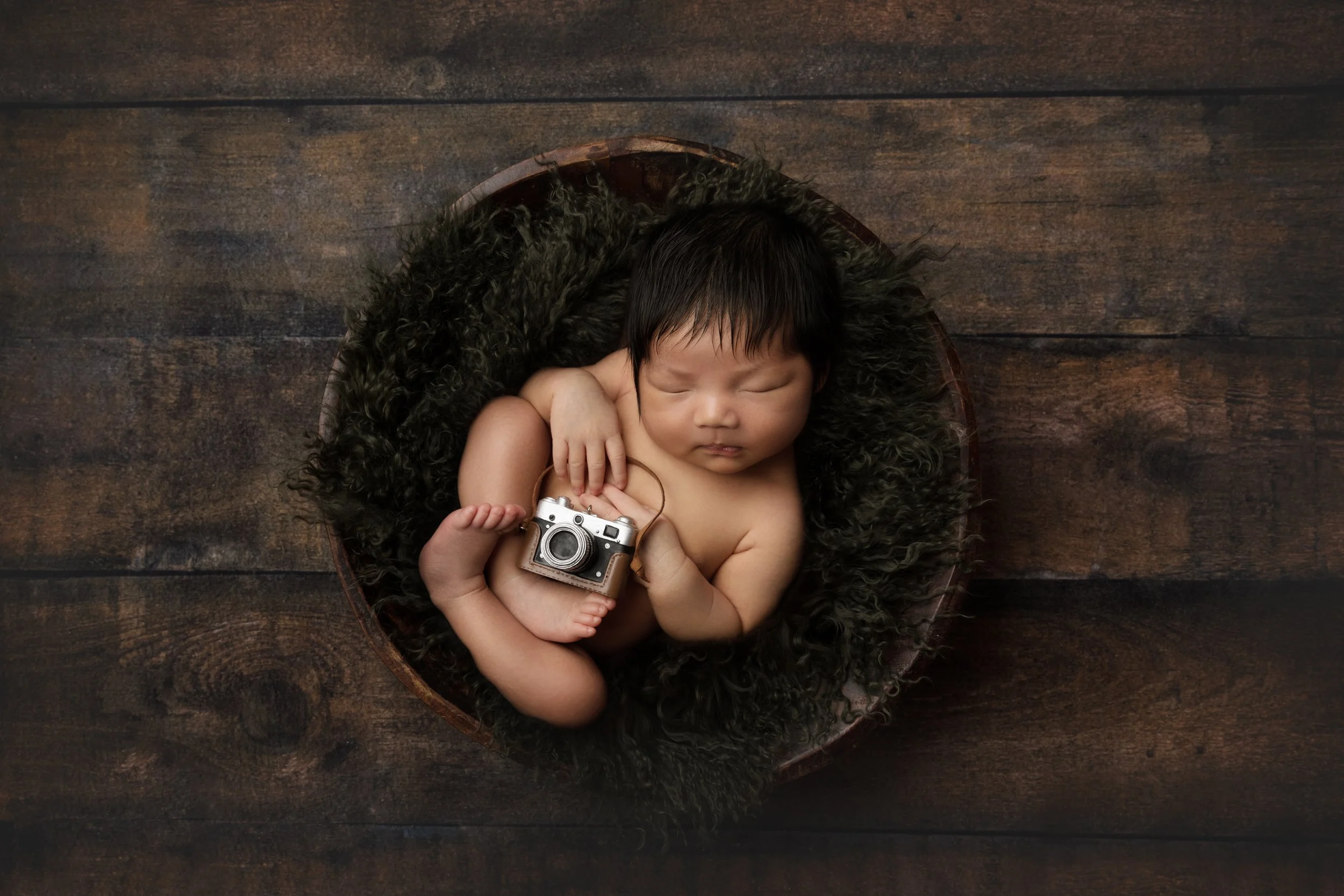 sleeping newborn baby boy curled up in basket with green fluff holding a tiny camera prop
