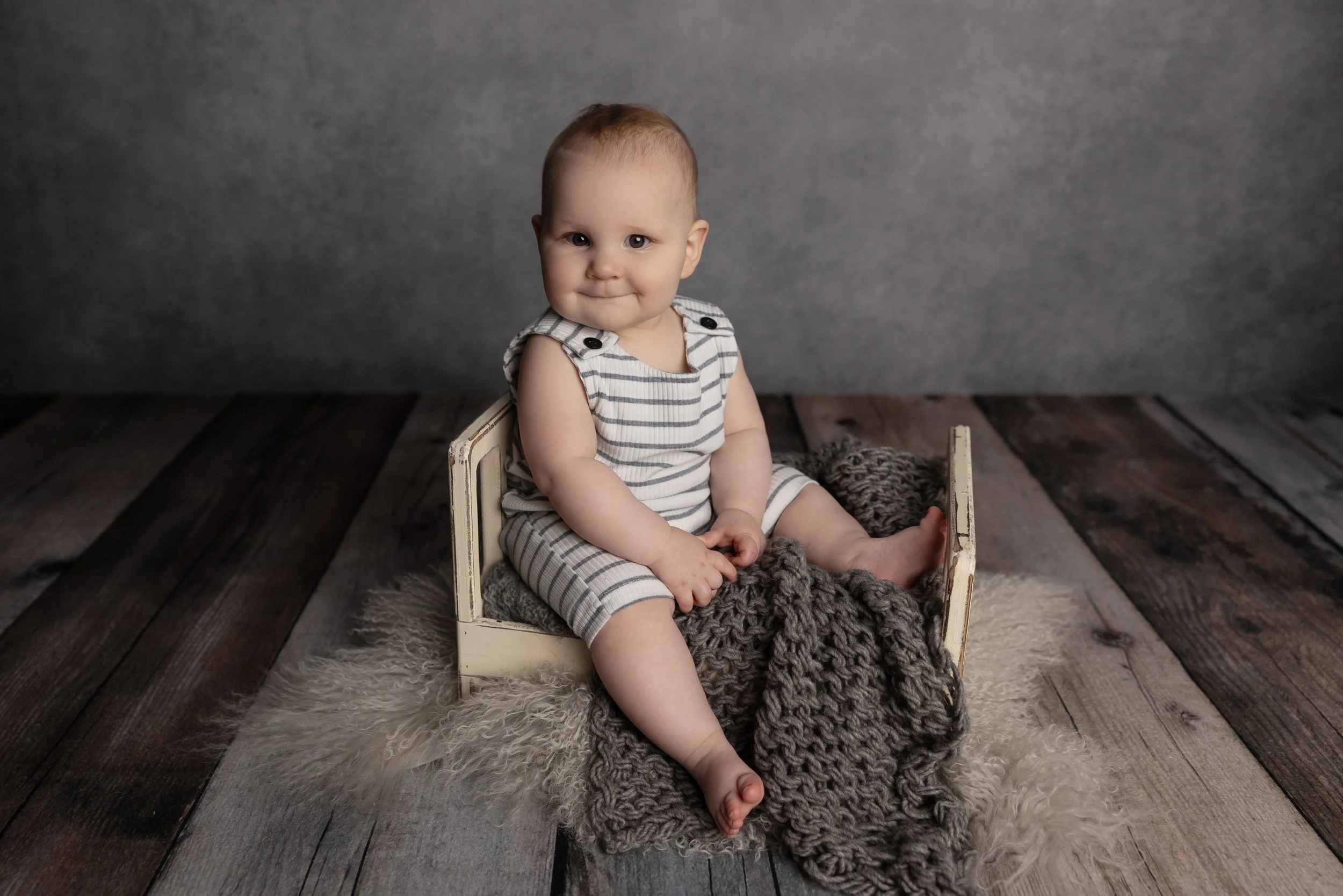 Eight-month milestone portrait of a baby sitting independently in the same miniature bed prop used during their newborn session.