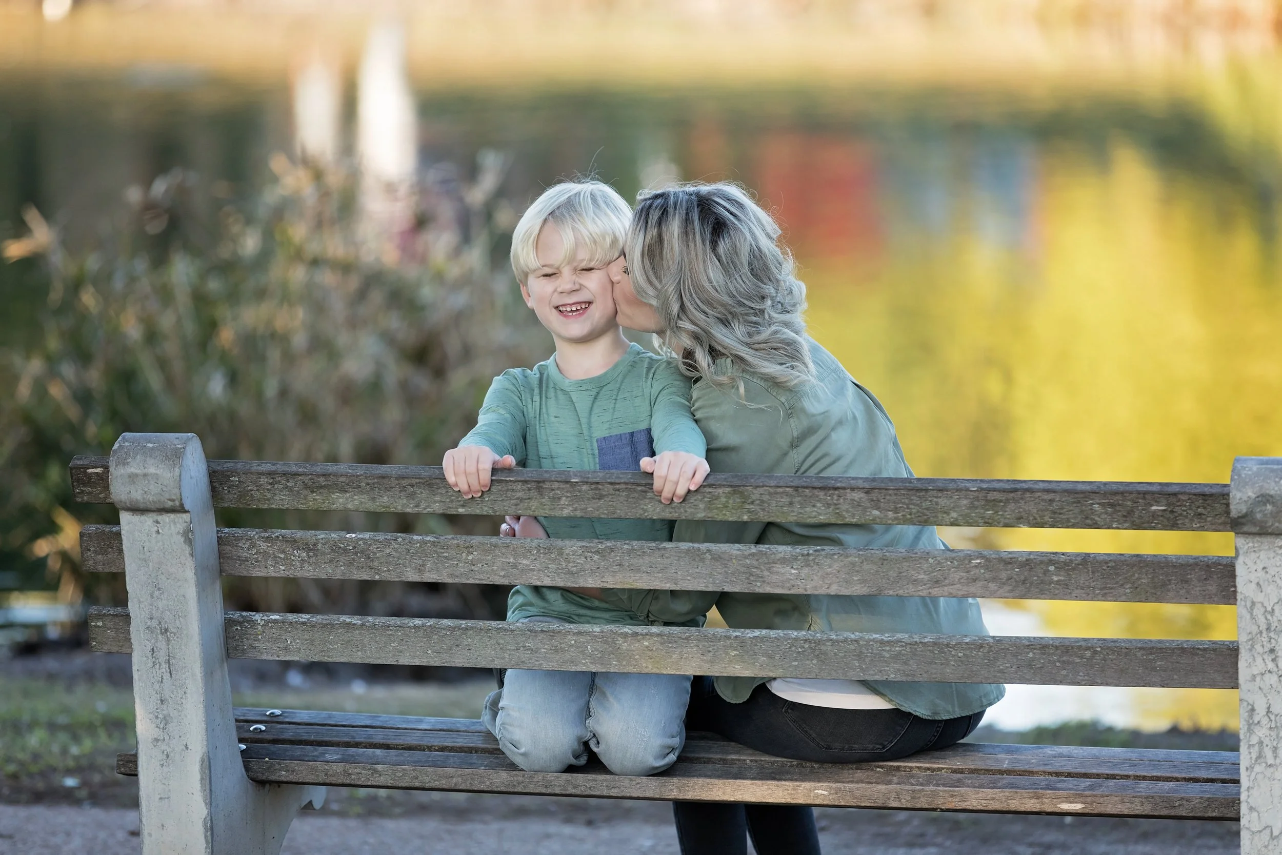 Mother kissing smiling son on a park bench during an outdoor family session