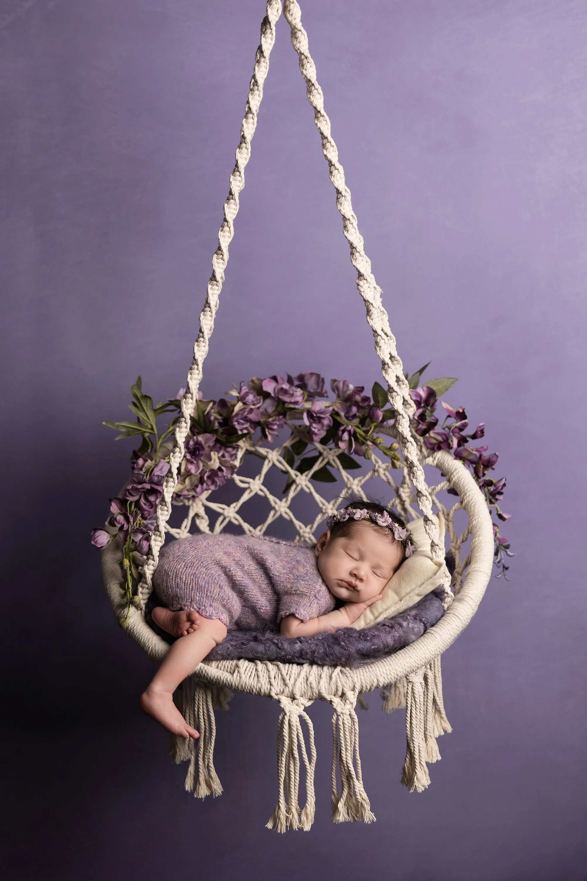 newborn baby girl sleeping in hanging swing with purple flowers photographed in Austin Texas photography studio