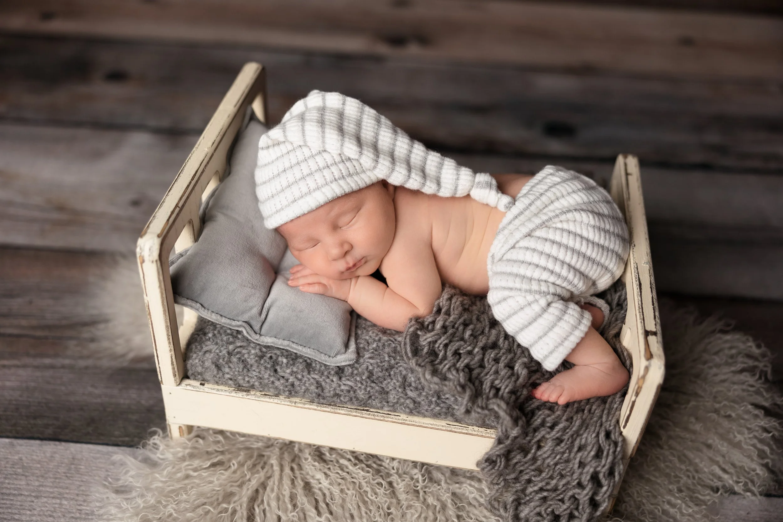 Newborn bed prop photography showing a sleeping baby in a classic bum-up pose with one tiny leg dangling over the edge of the bed.