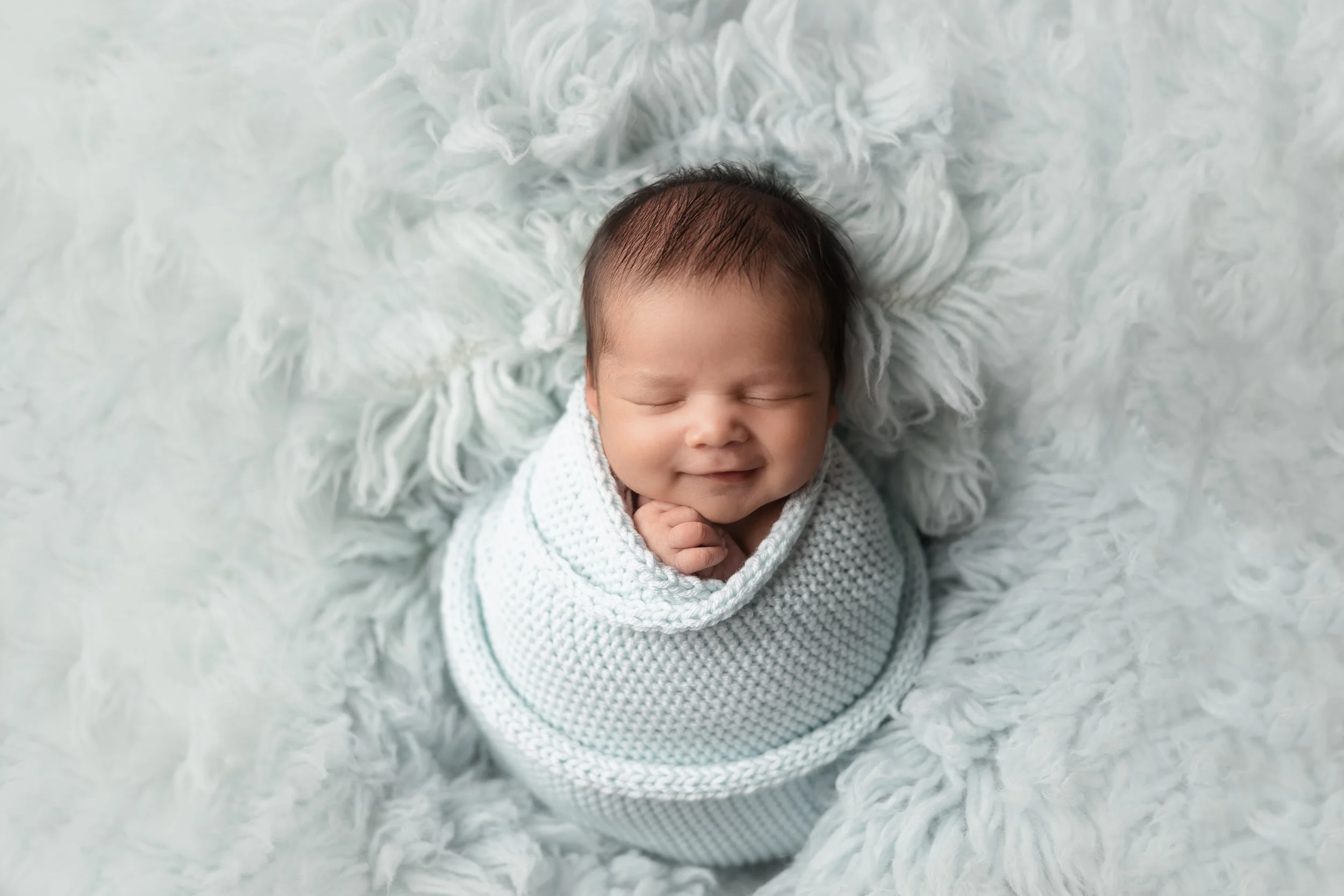 smiling newborn baby boy swaddled in a light blue wrap on a light blue rug