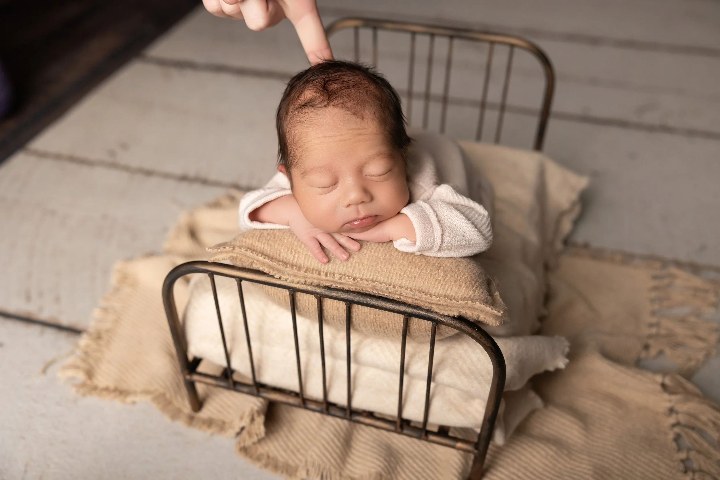 Behind-the-scenes newborn photography showing a parent or assistant spotting a sleeping baby while posing safely on a miniature bed prop.