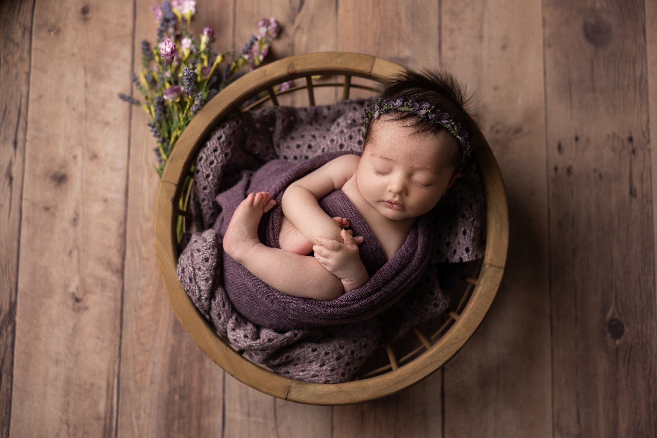 Posed newborn studio photo of baby wrapped and sleeping in a round wooden bowl with soft purple blankets and floral accents