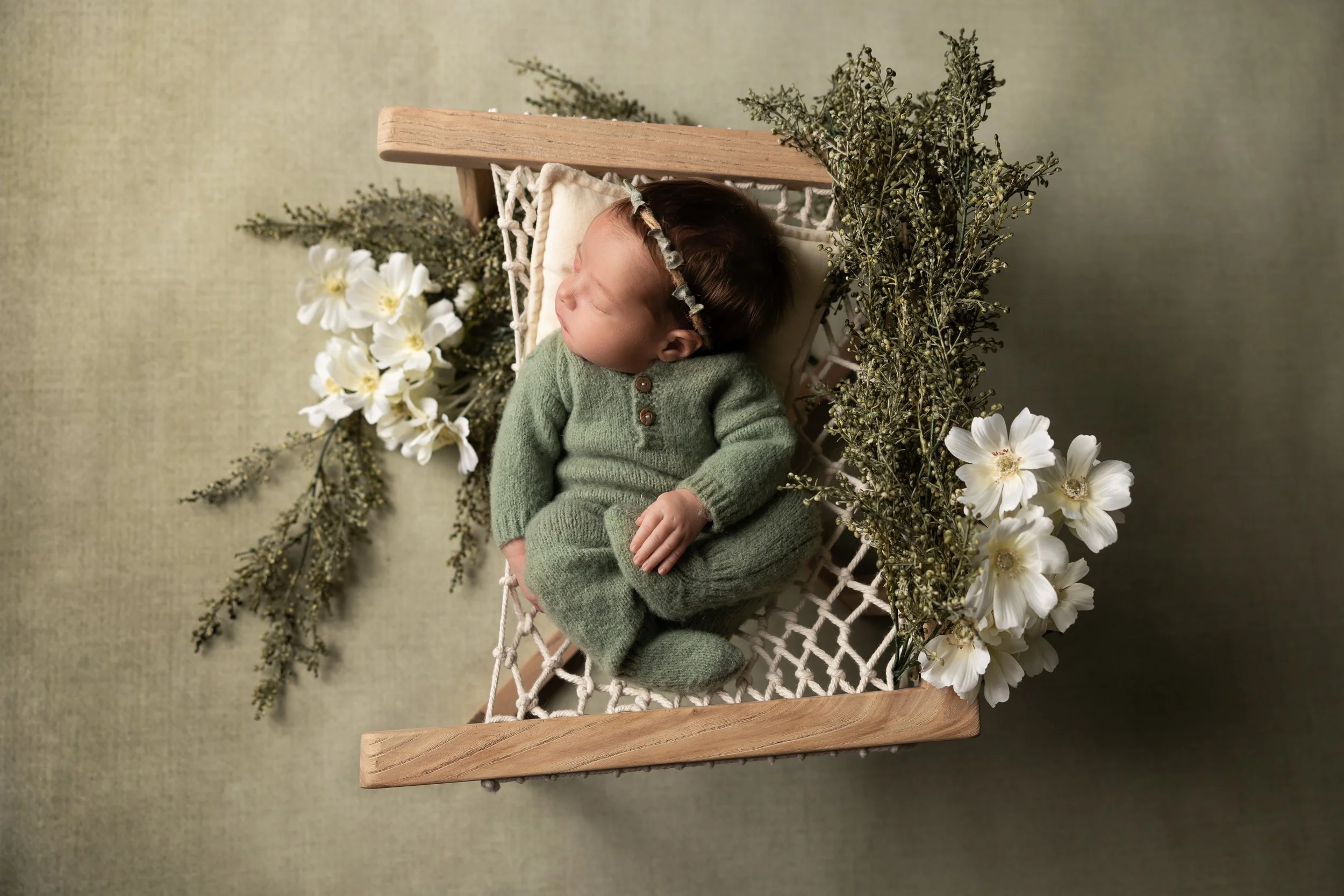 newborn girl wearing green romper posed sleeping and holding foot surrounded by greenery and flowers
