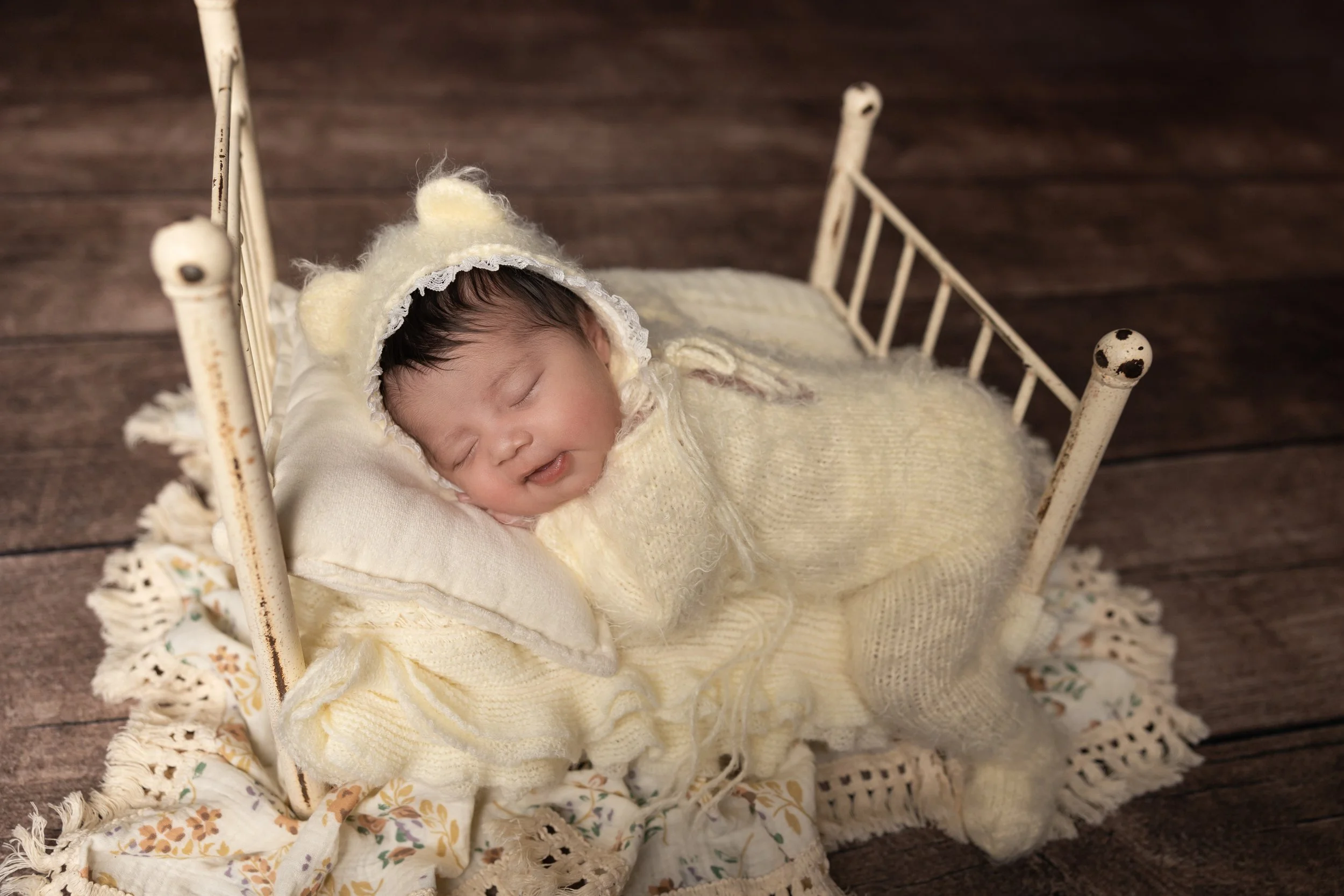Newborn bed prop photography featuring a baby in a cream bear bonnet posed in a classic bum-up position on a tiny bed with layered textures.