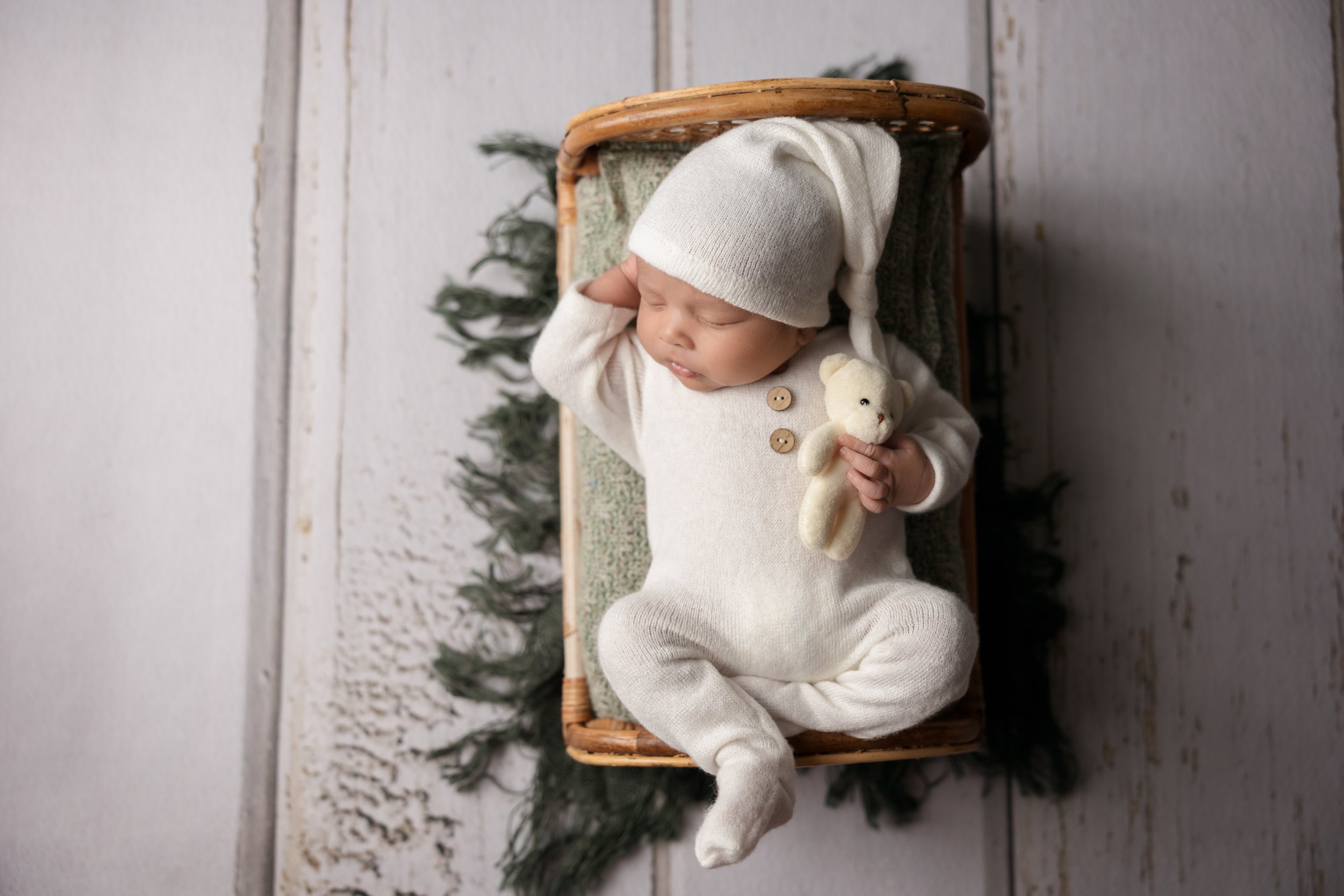 Austin newborn photography featuring a sleeping baby in a cream knit outfit and hat, posed in a basket holding a teddy bear in a relaxed newborn pose.