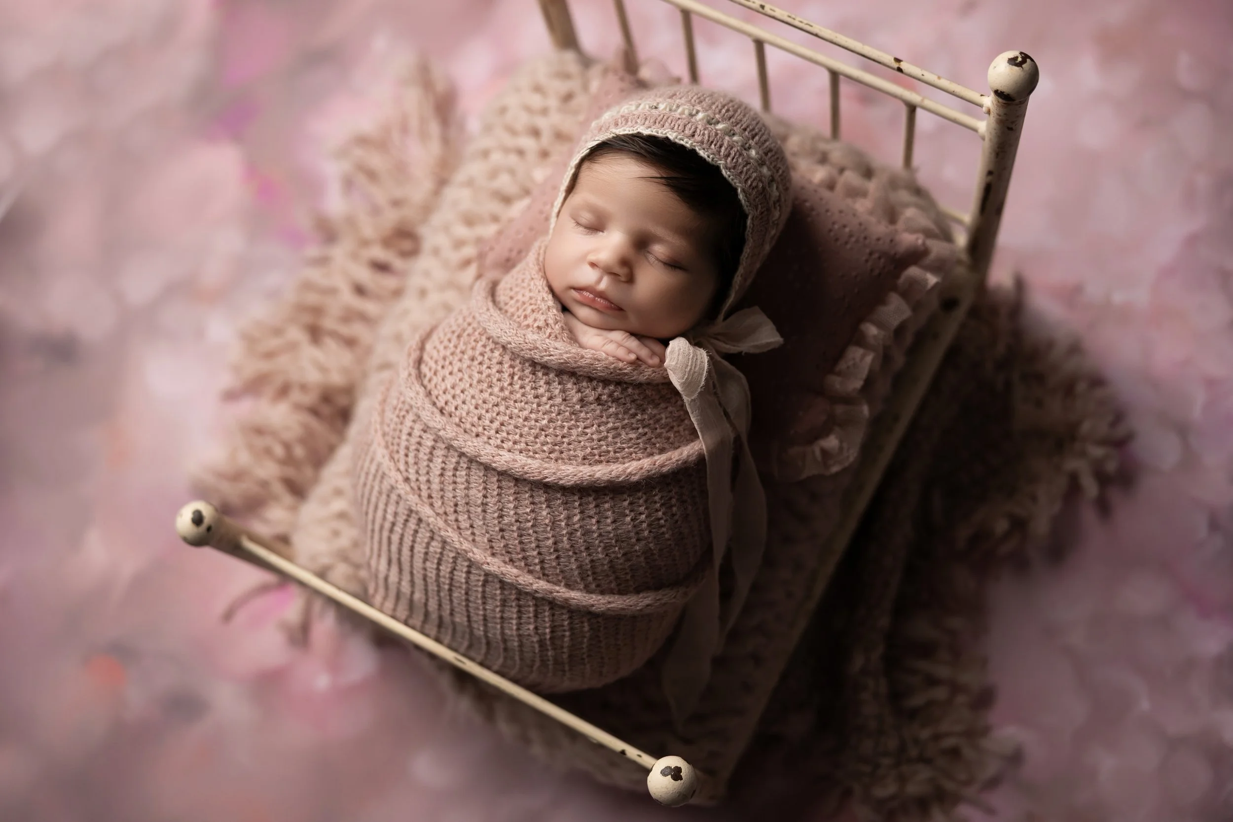 Angled newborn portrait of a sleeping baby wrapped in a blush swaddle on a miniature bed prop, demonstrating posing variety.
