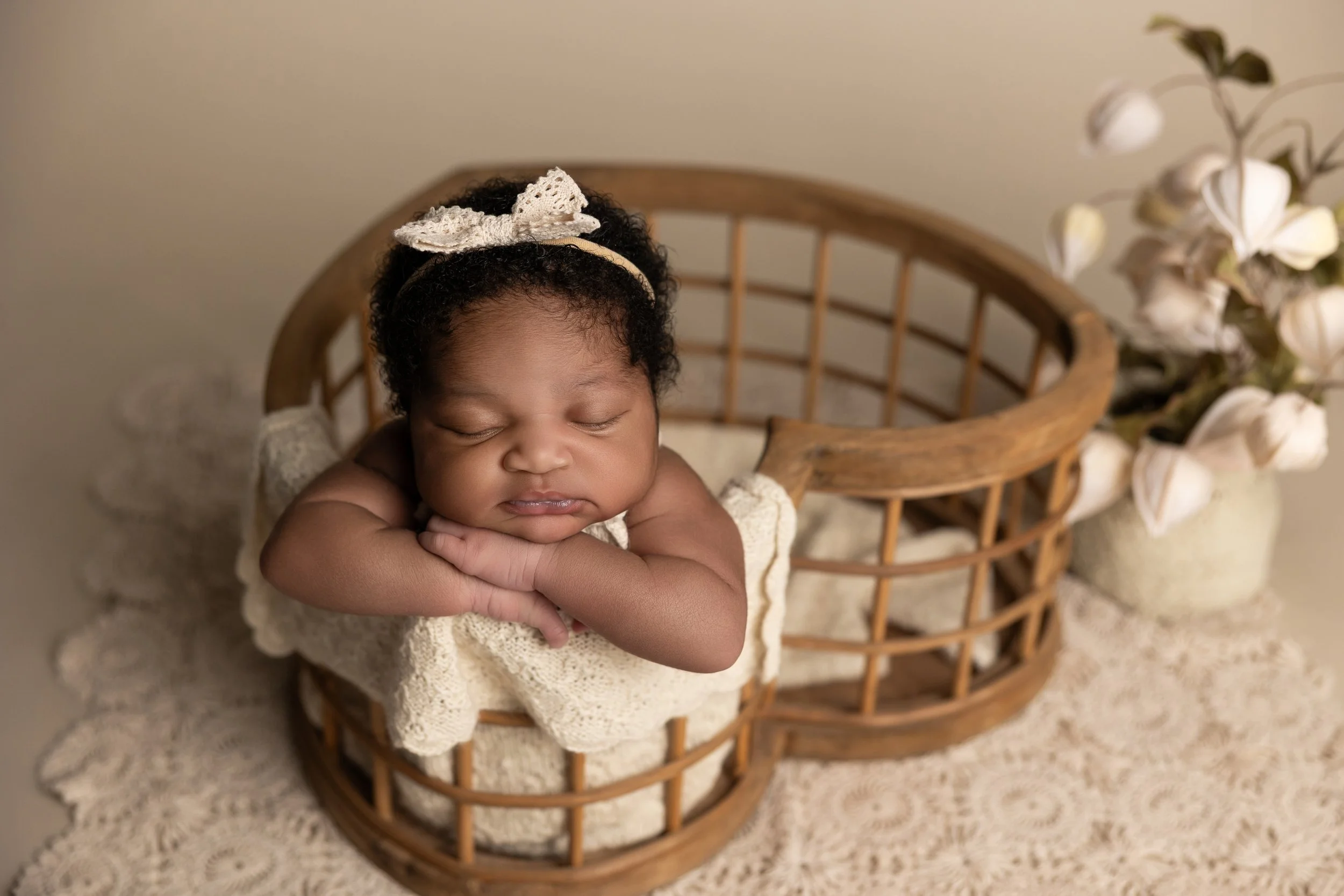 newborn girl posed with chin on hands in a heart shaped basket with lace and flowers