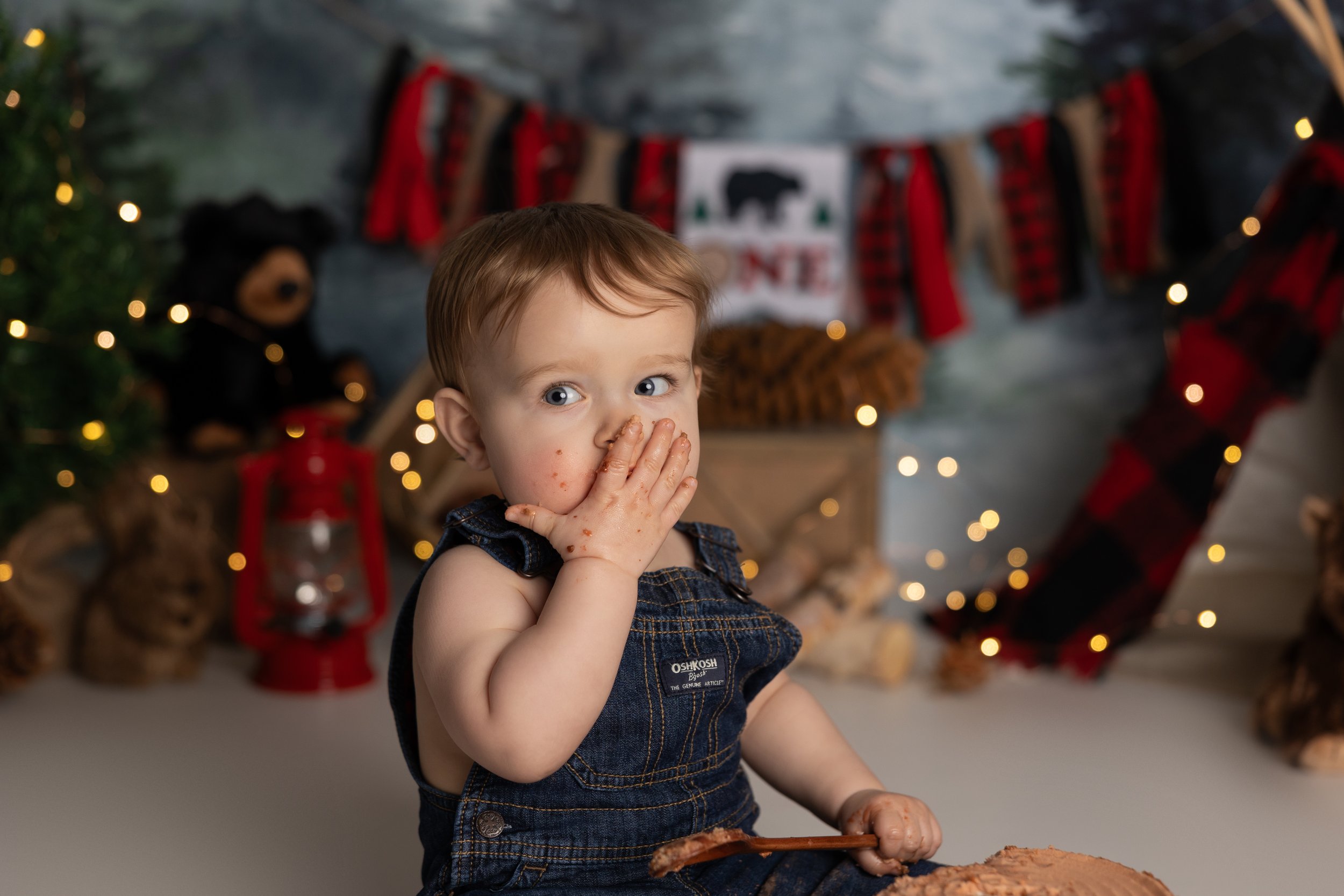 One year old boy in denim overalls eating cake during a rustic lumberjack-themed cake smash session in Austin Texas.