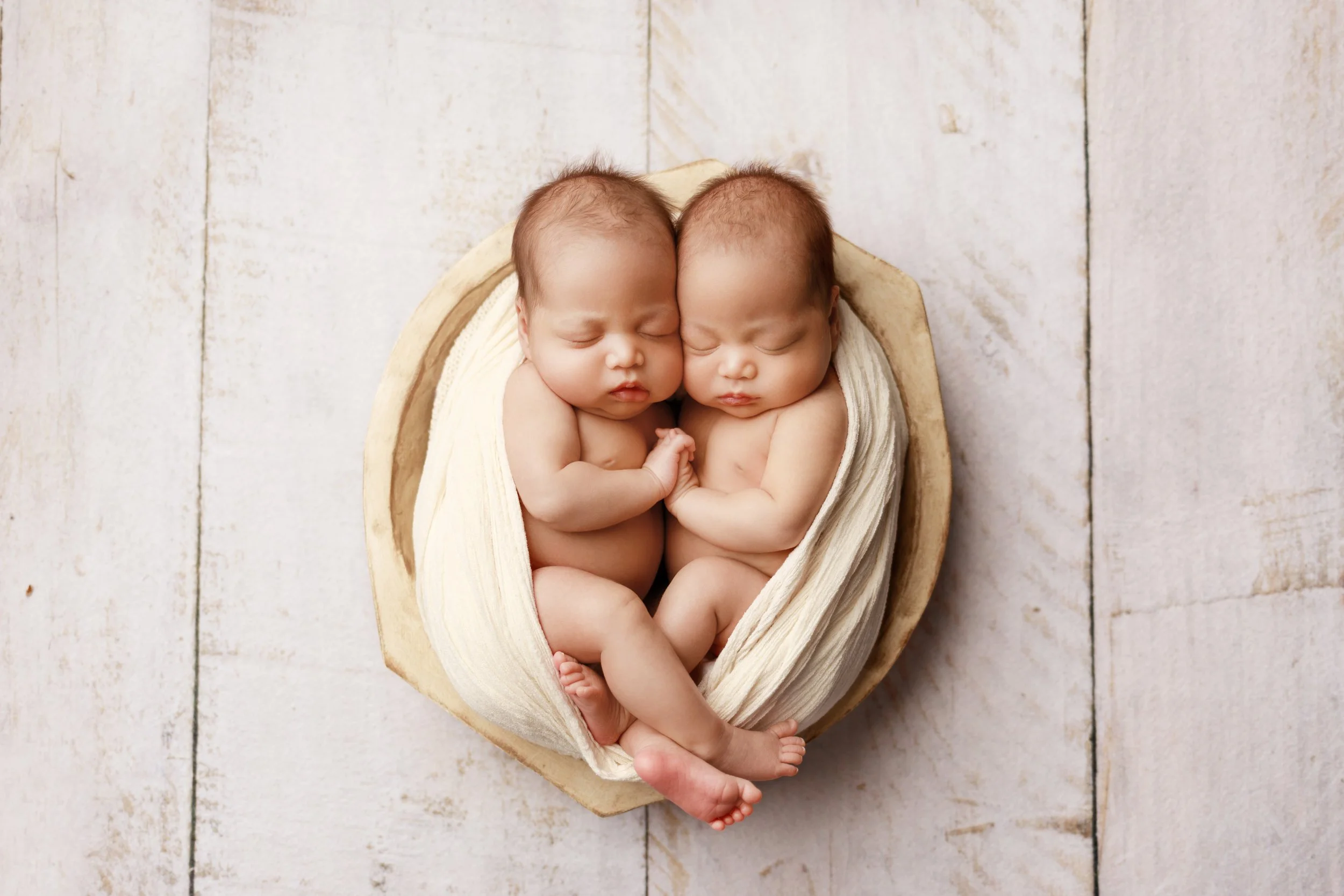 Twin newborn babies peacefully sleeping together in a soft cream wrap inside a wooden bowl during a posed studio newborn photography session in Austin, Texas.