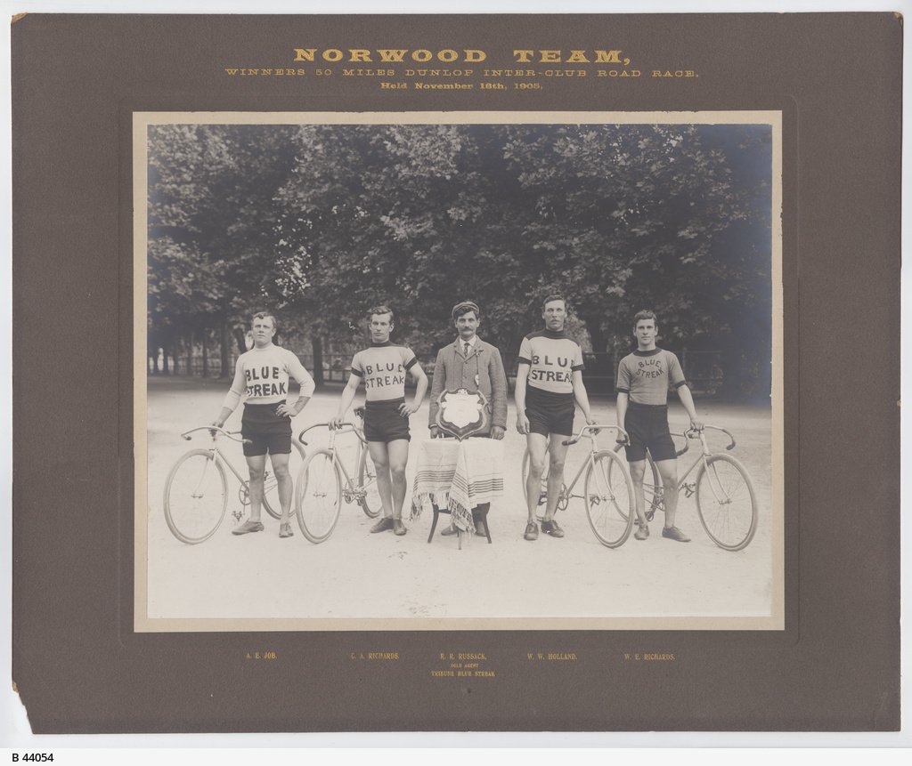 Members of the Norwood Cycling Club with their bicycles after winning the Dunlop 50 miles inter club road race; the wear shirts with 'Blue Streak' in large letters and stand by a small table holding the winning shield 1905