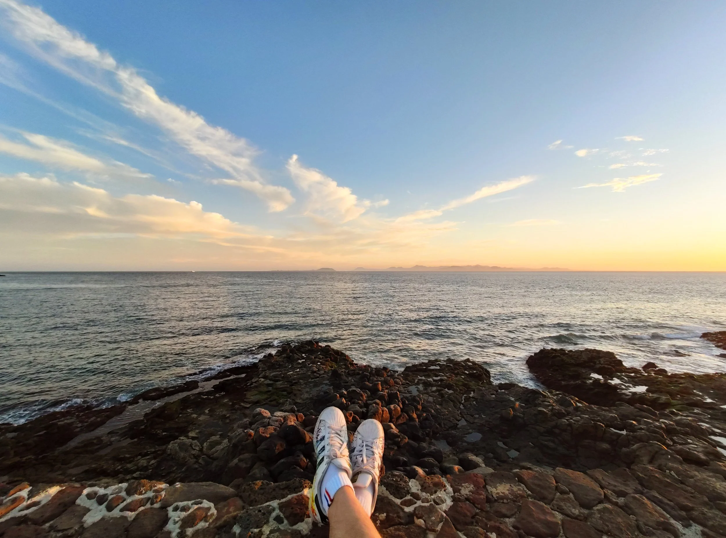 Person lying on rocky shore, wearing white sneakers, looking out over the ocean during sunset with a colorful sky and distant landforms.