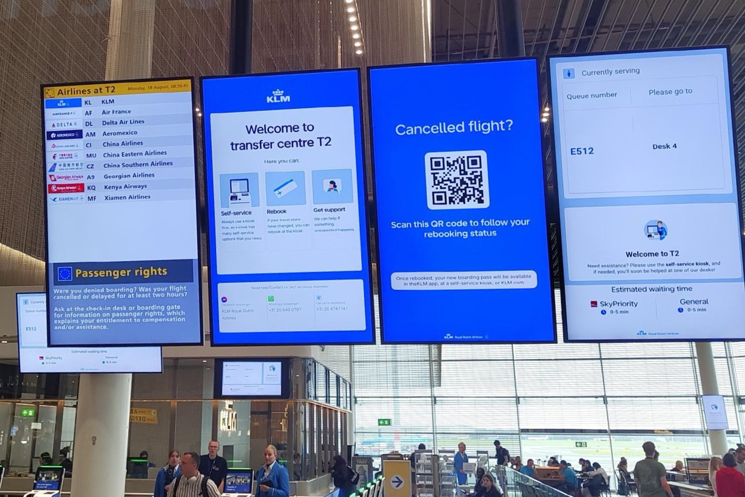 KLM disruption screen at Schiphol Transfer Centre displaying a QR code for passengers to follow their rebooking status, shown alongside the regular airport information screens