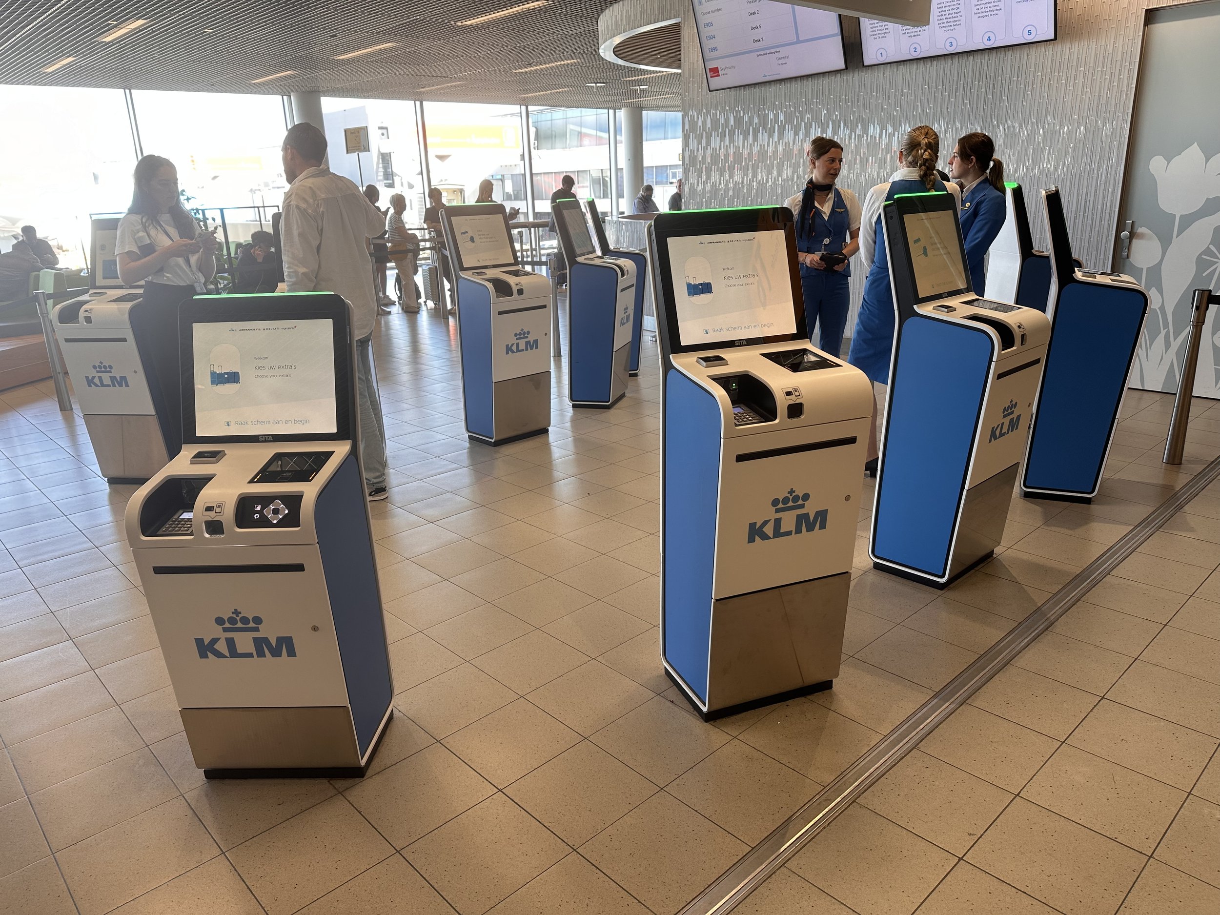 Row of KLM self-service kiosks with airport agents assisting passengers at Schiphol