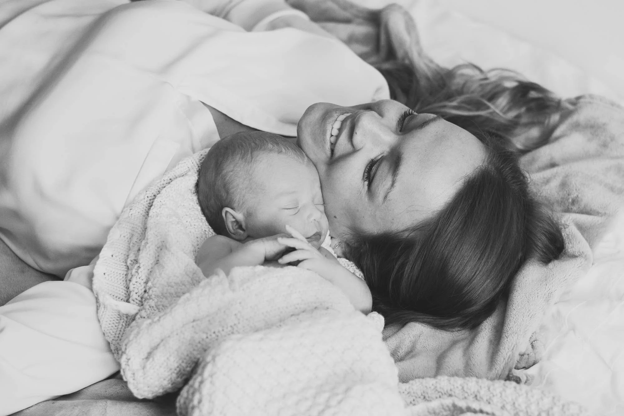 Zwart-wit foto van een vrouw die op bed ligt met haar hoofd op een kussen en in haar armen een slapende baby, beiden glimlachend.