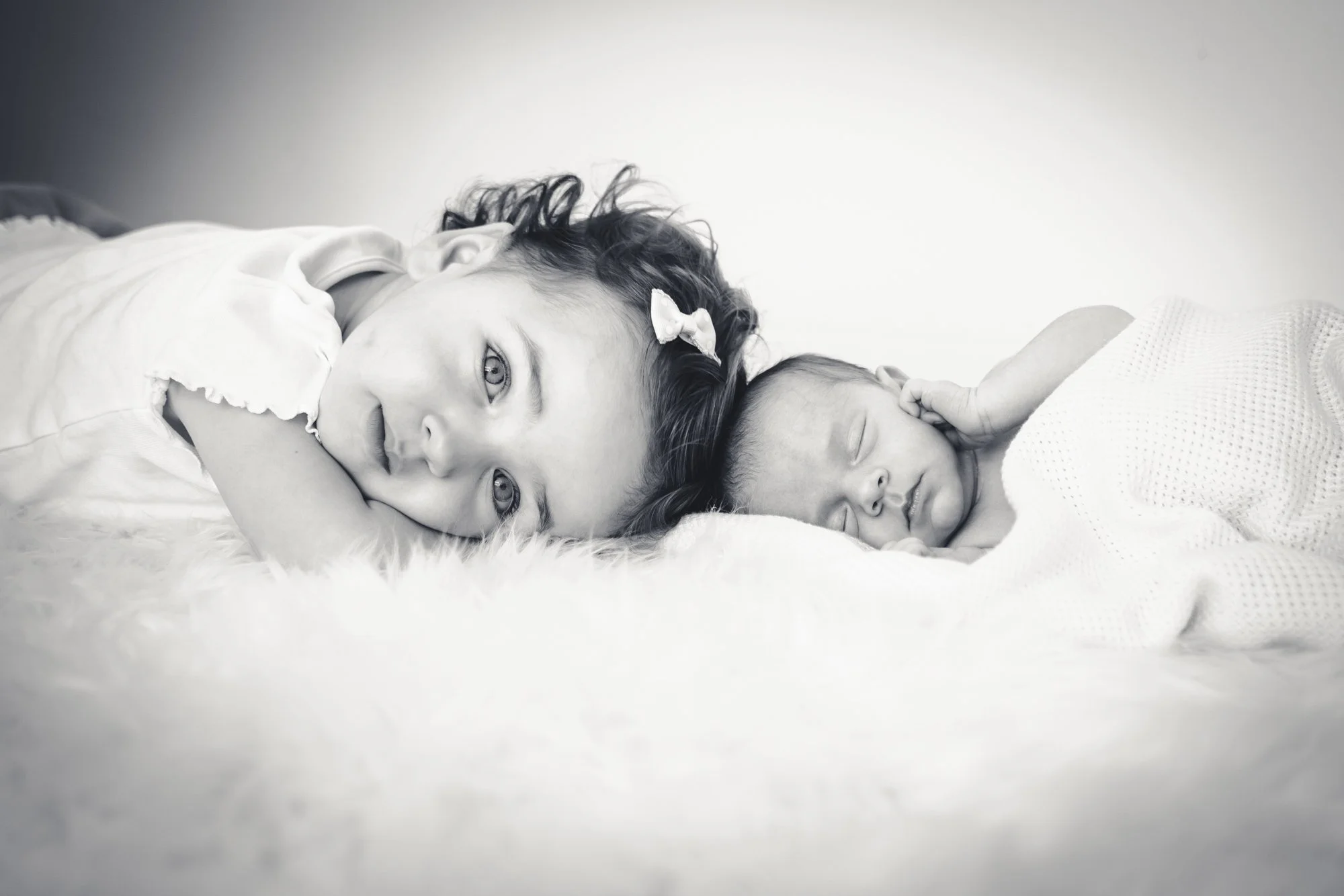 Zwart-wit foto van een jonge vrouw en een baby die samen op een bed liggen. De vrouw ligt op haar zij en kijkt naar de camera, terwijl de baby naast haar slaapt.