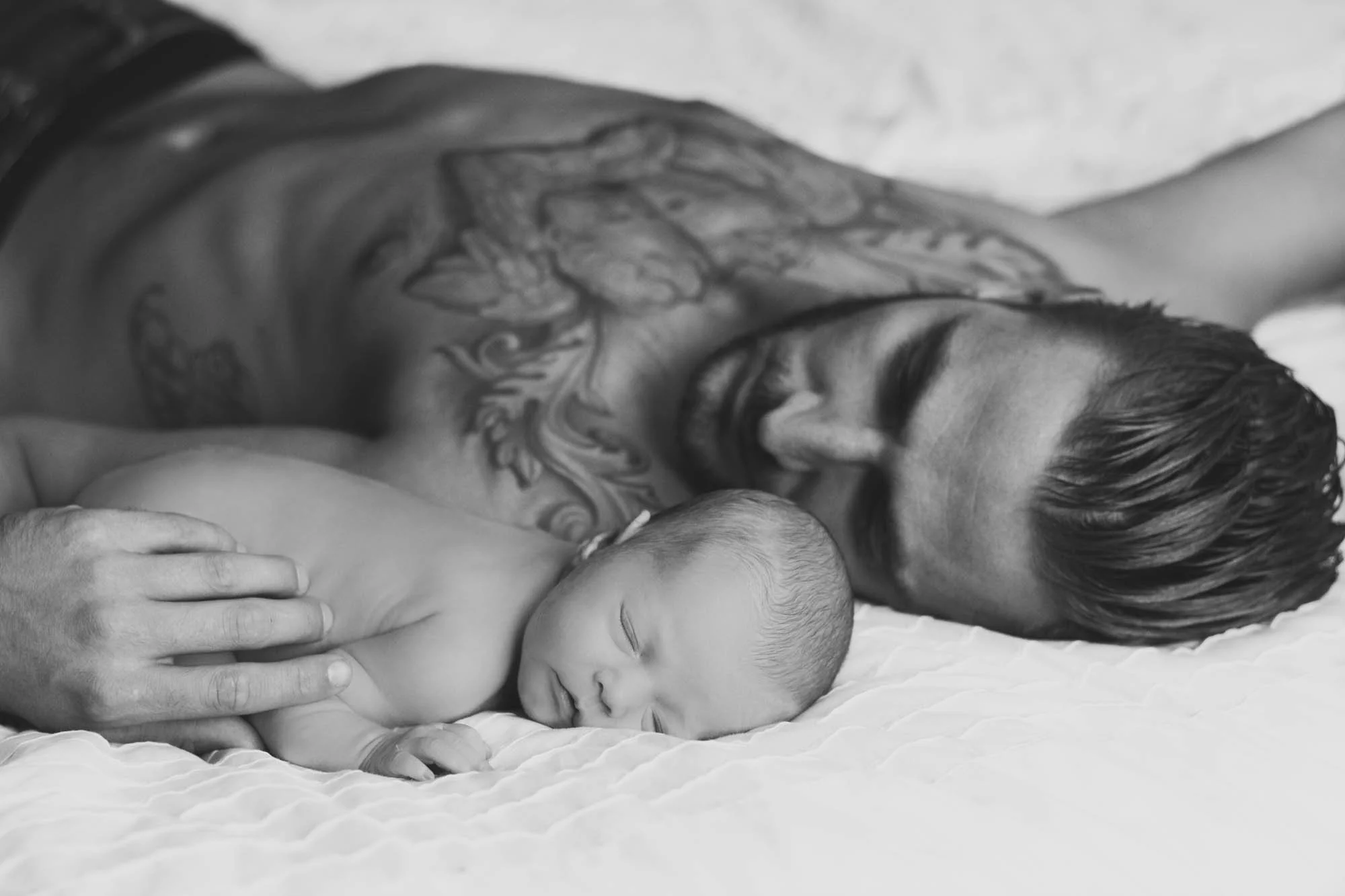 Zwart en wit foto van een lachende man met tattoos die een slapende baby vasthoudt