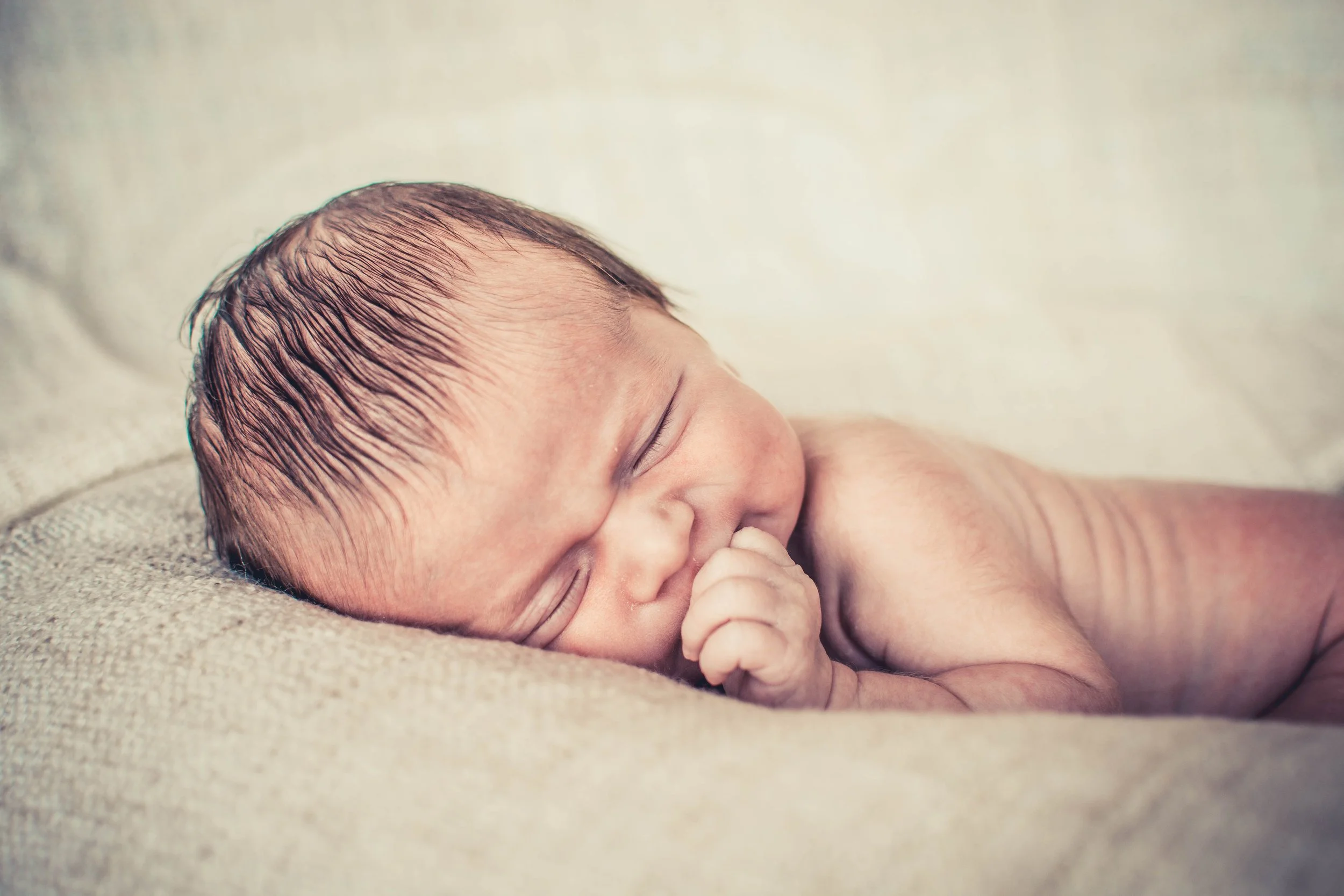 Een pasgeboren baby ligt rustig op een zachte, lichte ondergrond, met gesloten ogen en een kleine hand die naar de mond mensen. Het haar is nat en de huid is rozeachtig.