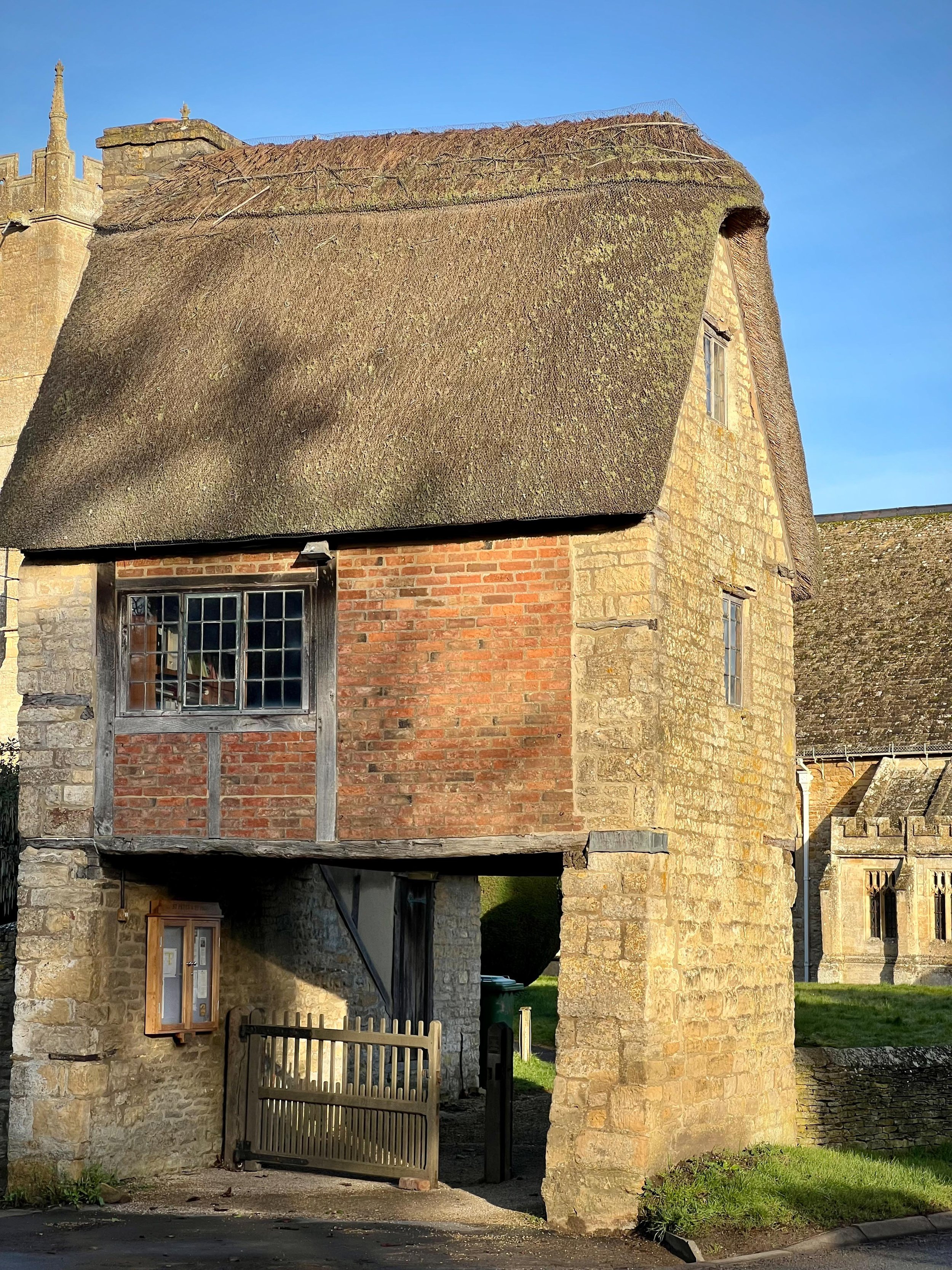 Historic stone building with a thatched roof, an archway with a wooden gate at the bottom, and a wooden-framed window on the side.
