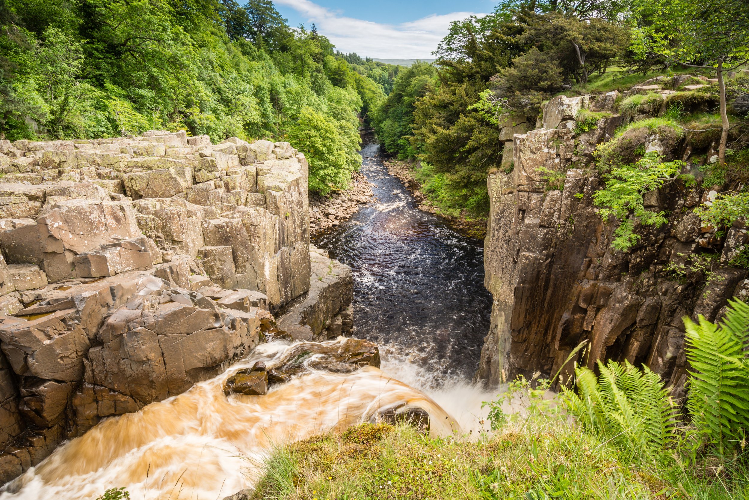 High Force waterfall scene with rocky cliffs and river in lush green forest, County Durham, England.