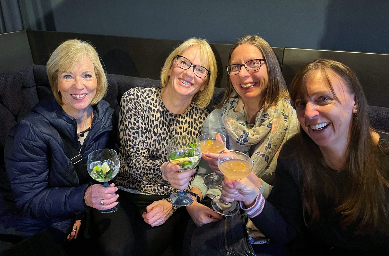 Four women smiling and holding drinks in a cozy setting.