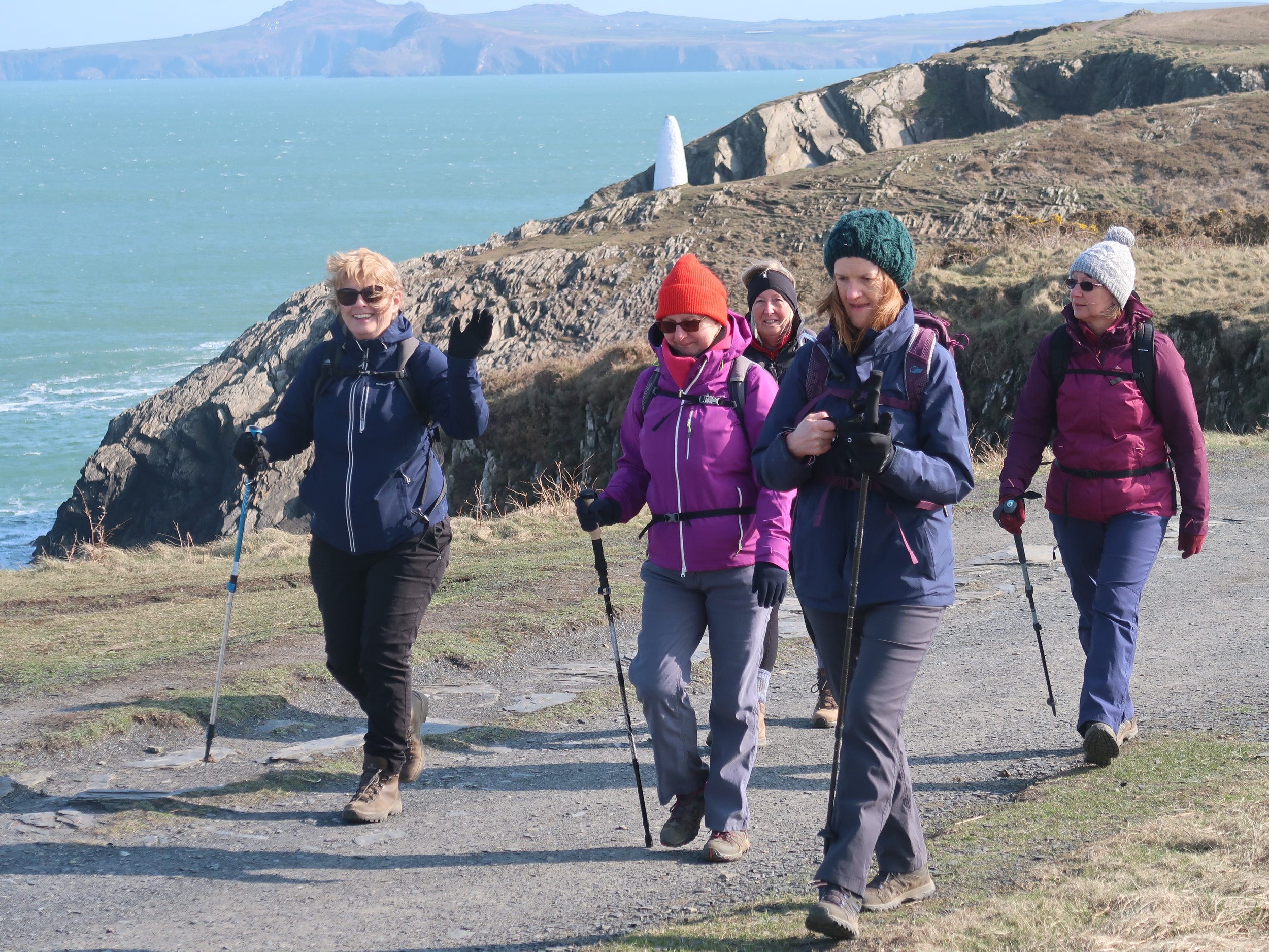 Group of women hiking along a coastal trail with walking poles, rocky cliffs, and ocean in the background.