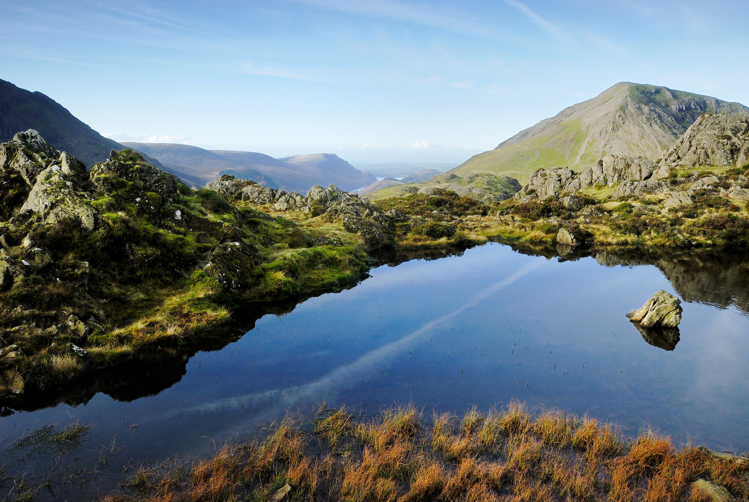 Scenic mountain landscape with a rocky pond, grassy slopes, and distant peaks under a clear blue sky.