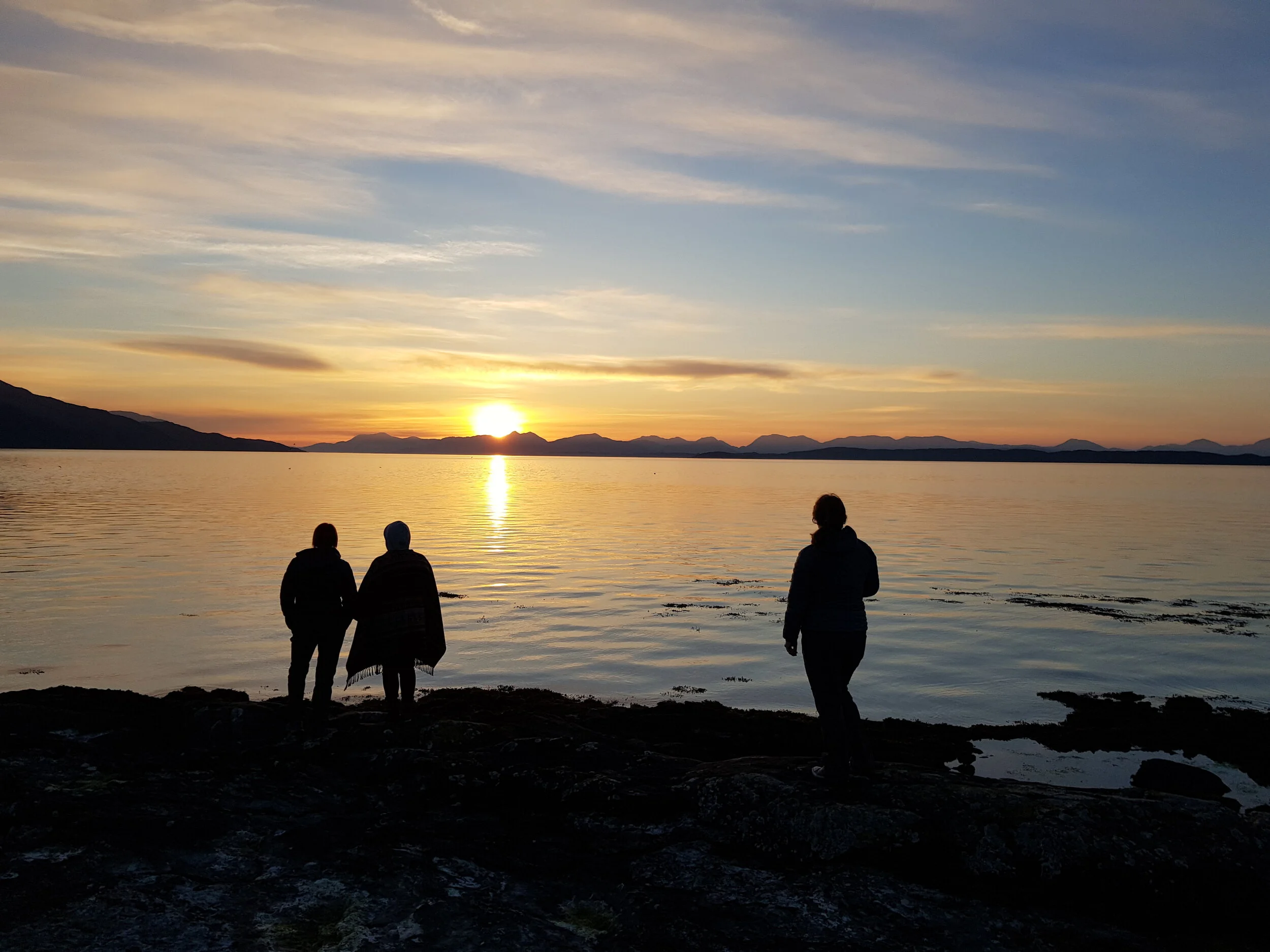 Three people silhouetted against a sunset over a calm sea and distant mountains.