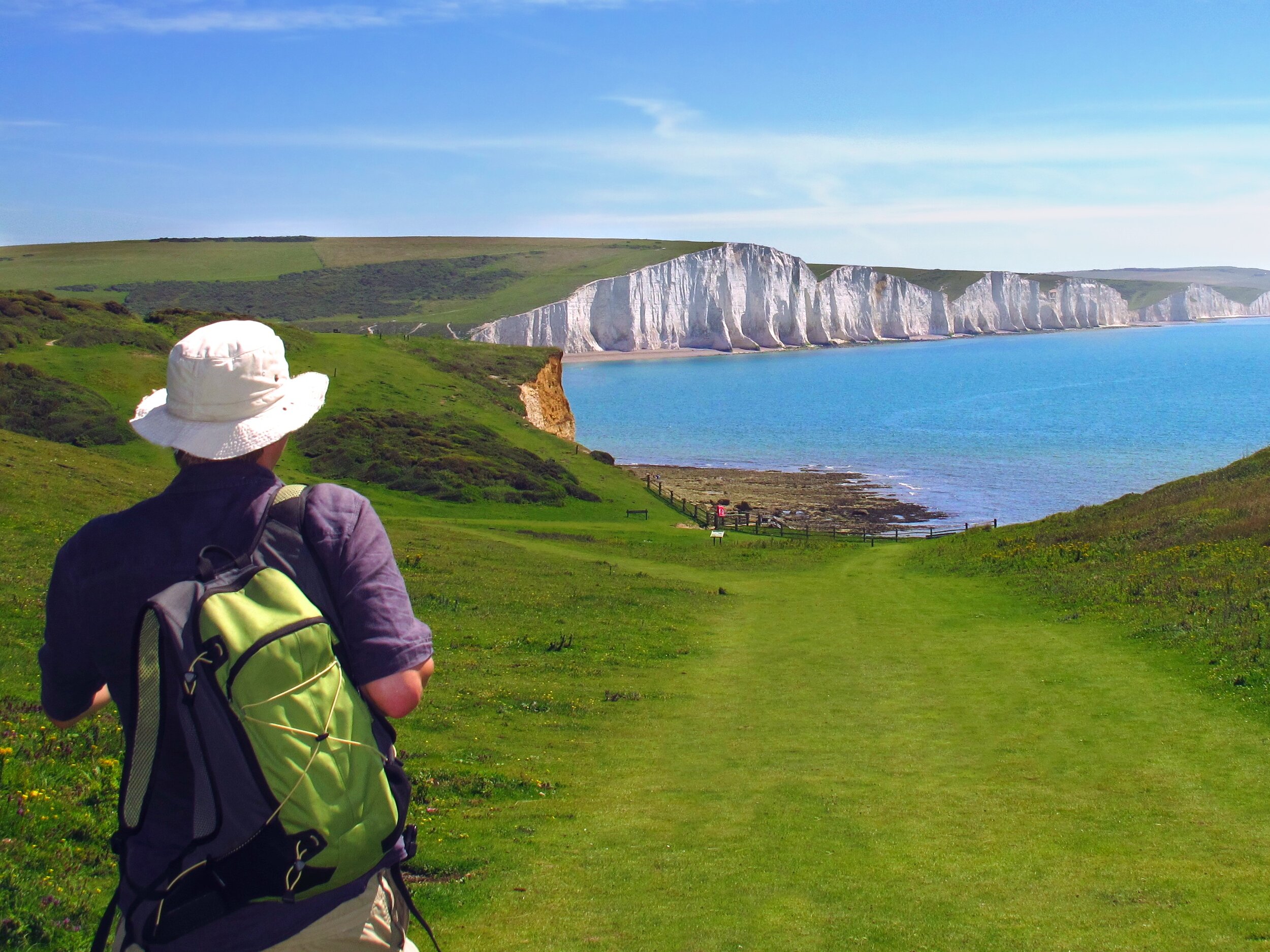Person with backpack walking on grassy path towards white chalk cliffs by the sea.