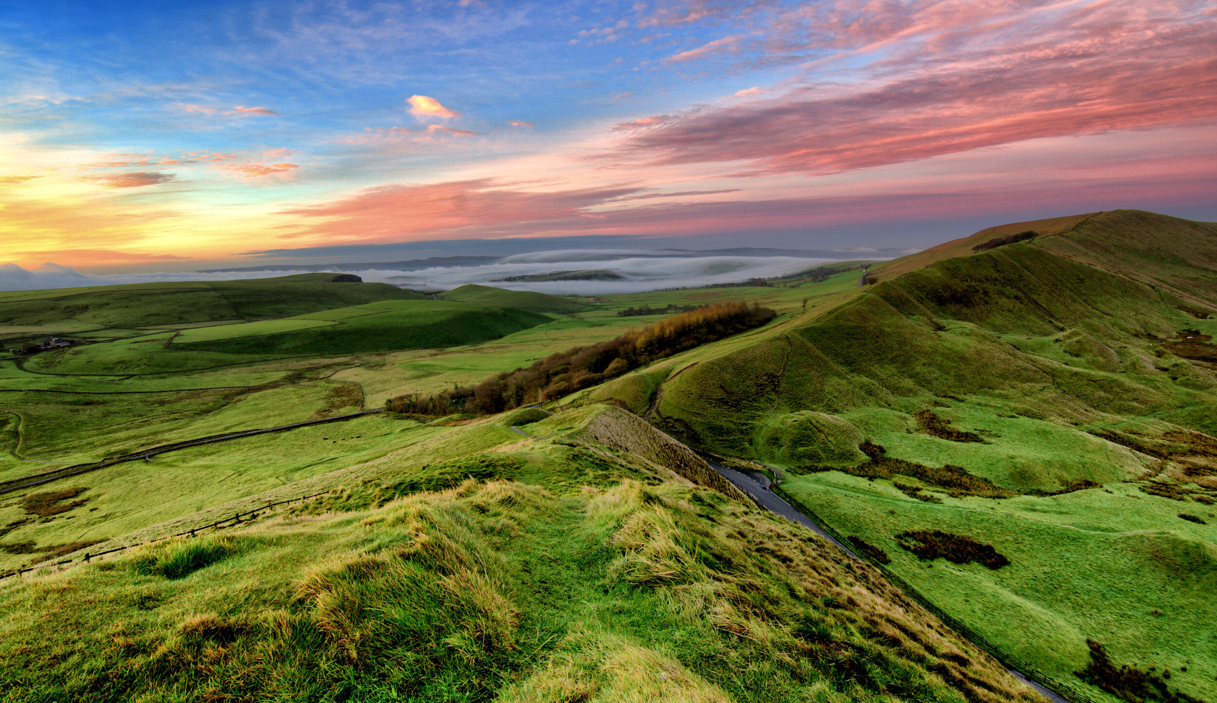 Rolling green hills under a colorful sky at sunset with scattered clouds, showcasing a scenic landscape view.