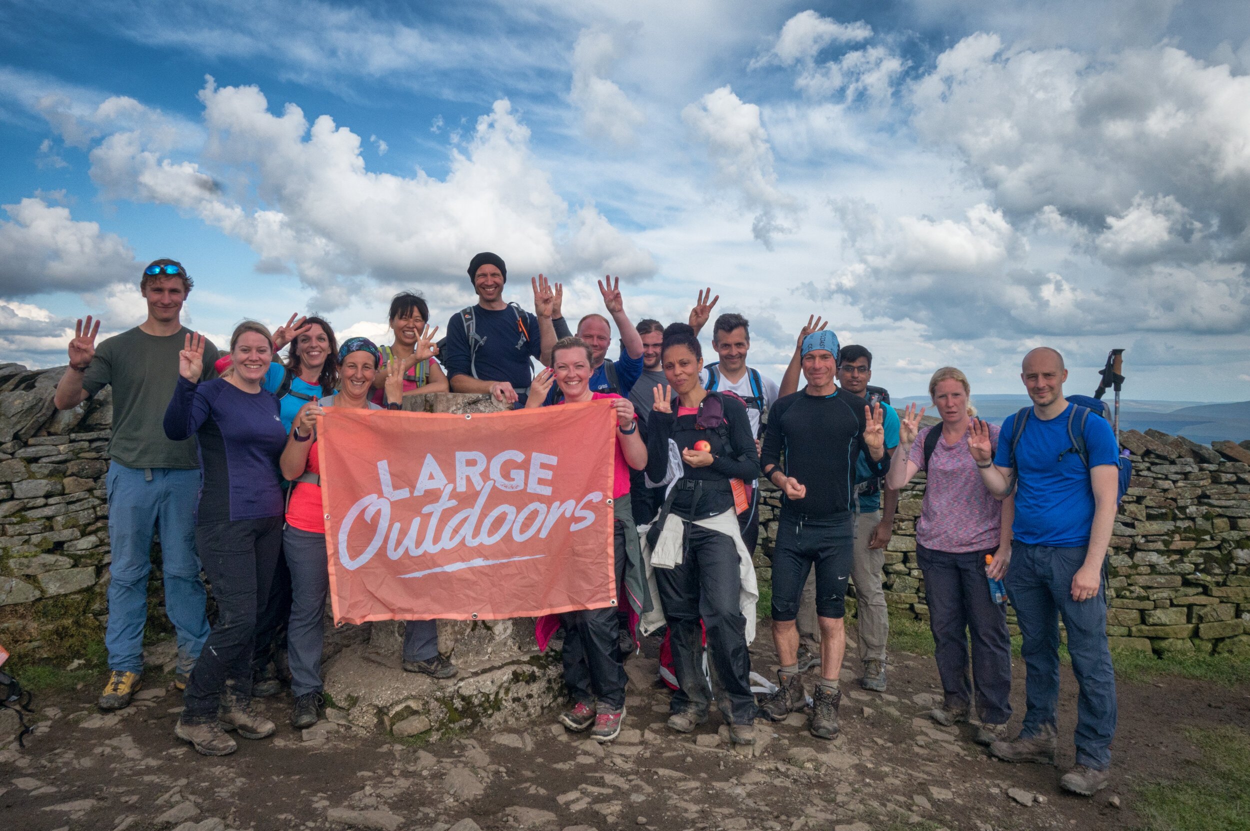 Group of hikers holding a "Large Outdoors" banner on a hilltop under a cloudy sky.