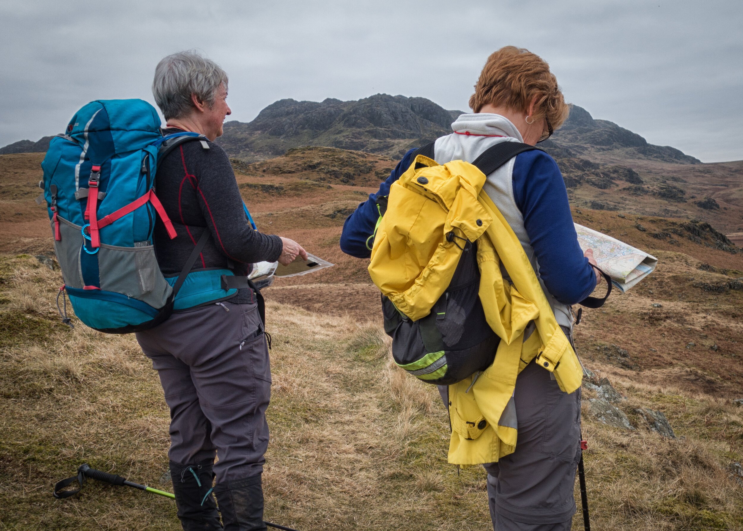 Two people wearing backpacks, consulting maps on a hiking trail in a rocky, mountainous landscape.