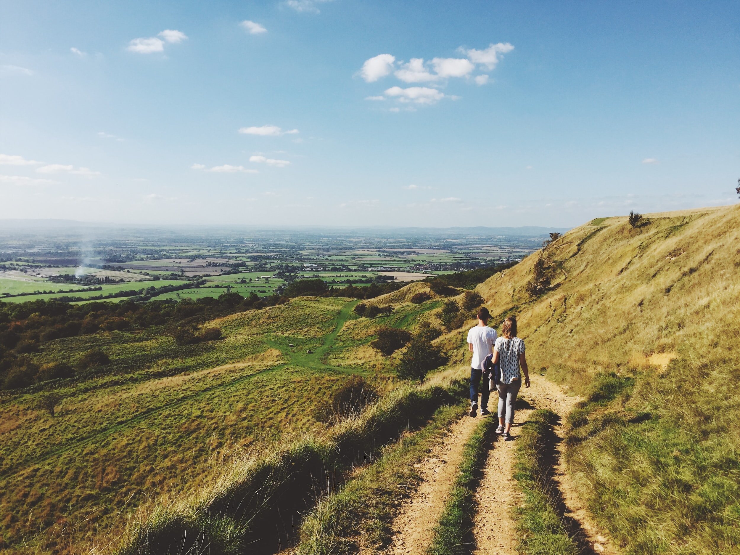 Two people walking on a grassy hillside trail overlooking a vast, scenic landscape with fields and a distant horizon under a clear blue sky.