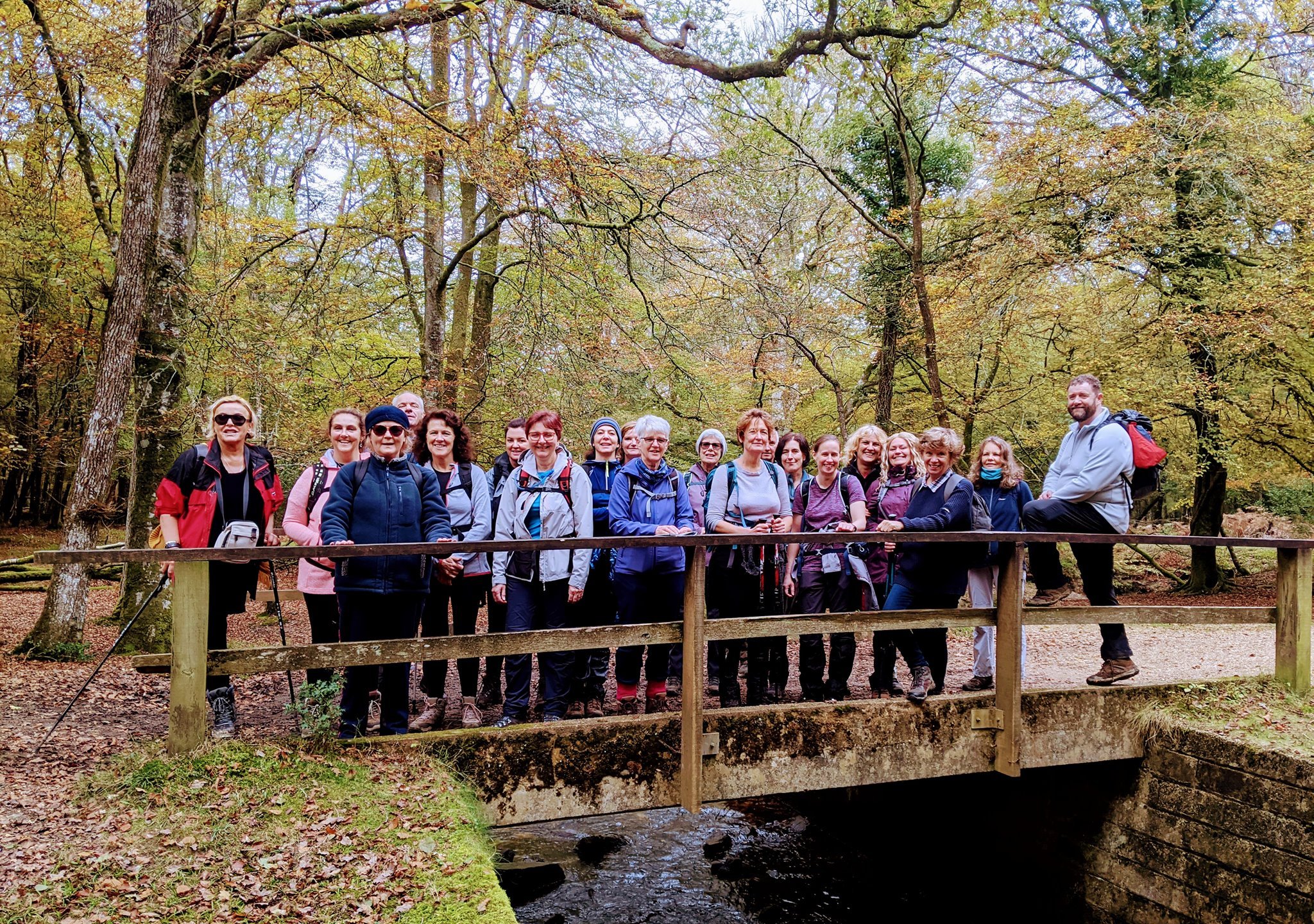 Group of people standing on a wooden bridge in a forest during autumn, wearing casual outdoor clothing.