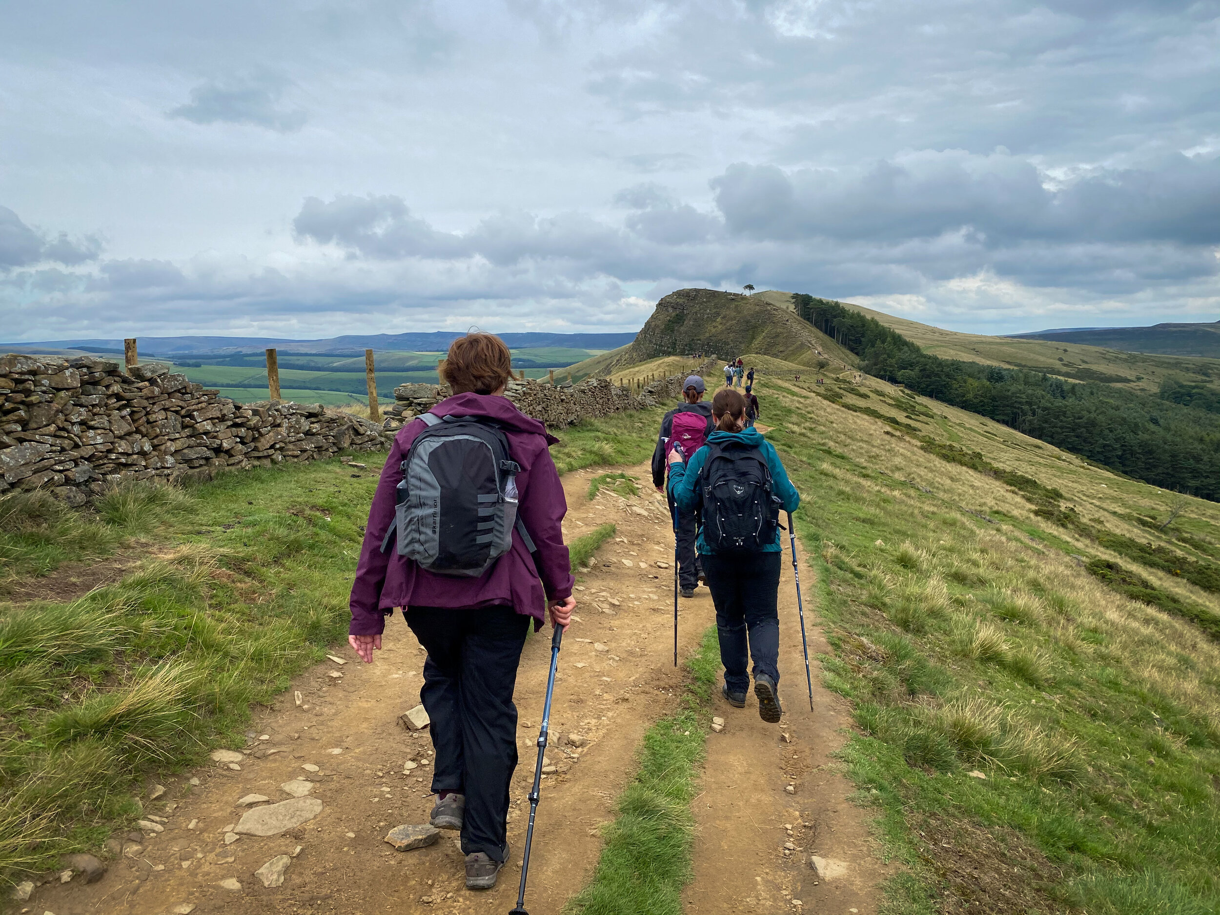Group of hikers walking on a dirt path near a stone wall and grassy hills with hiking poles and backpacks.