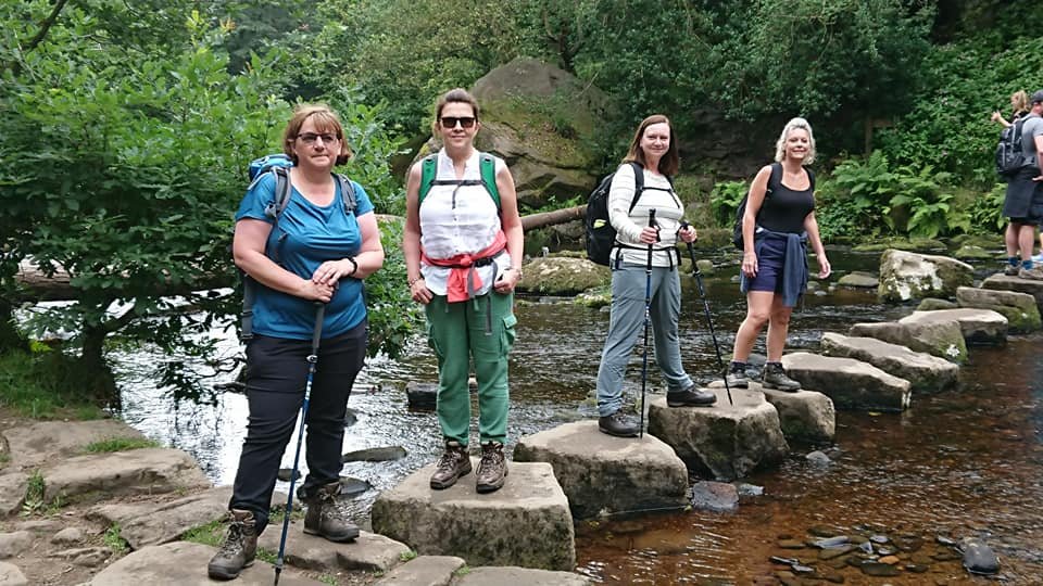Four women hiking on stepping stones across a shallow stream with lush greenery in the background.