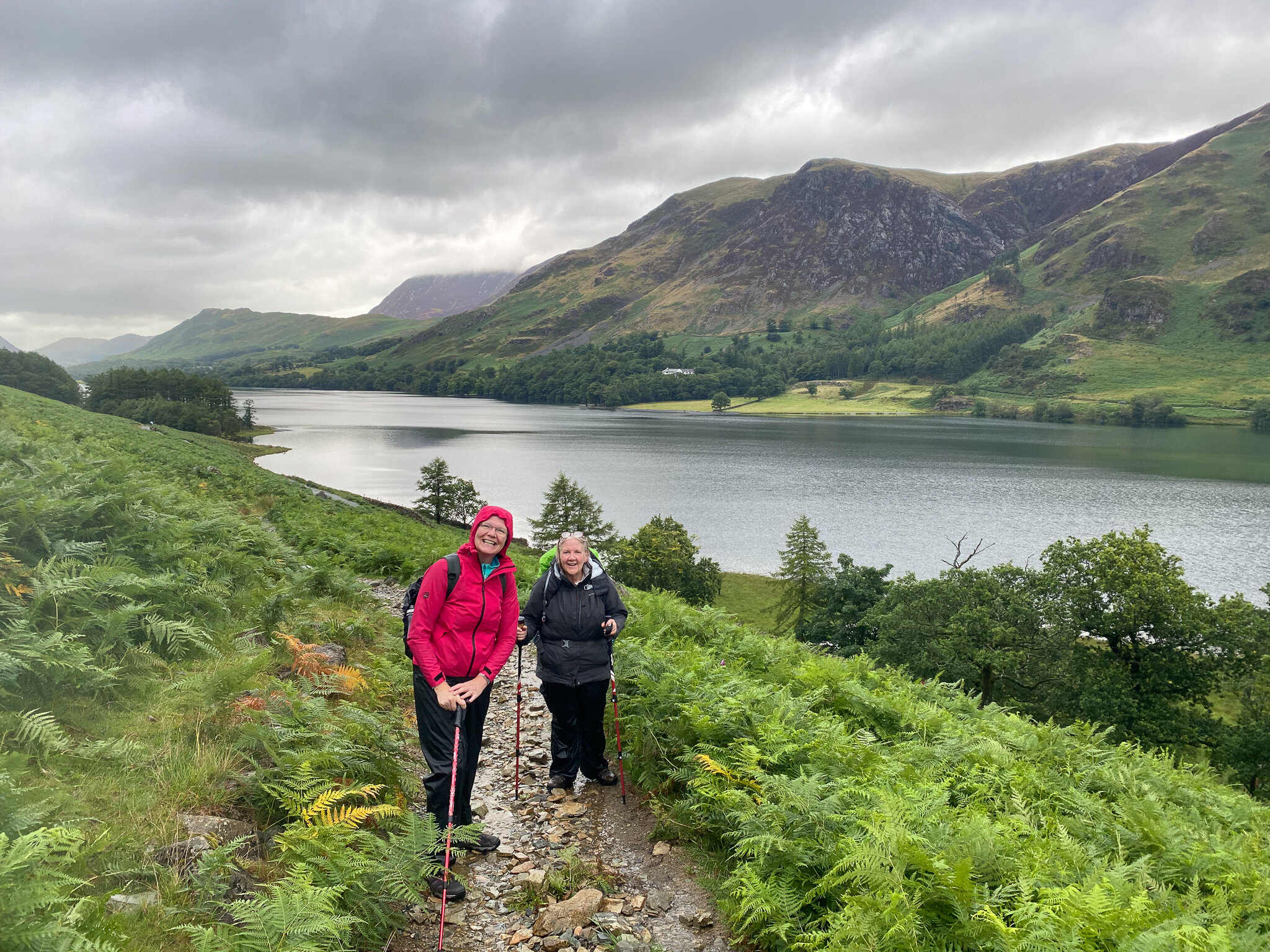 Two people hiking on a trail overlooking a lake and lush green hills, under a cloudy sky.