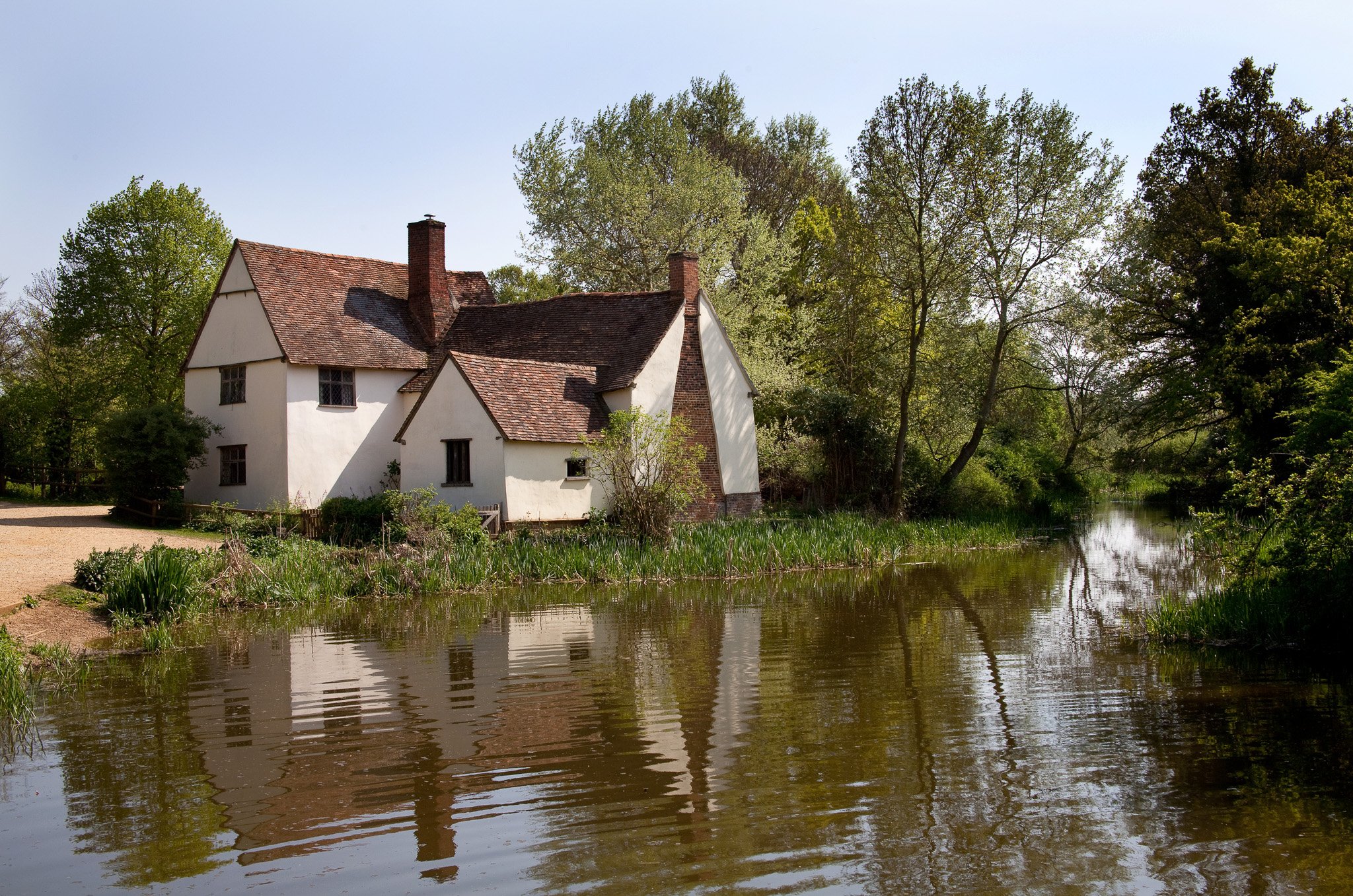 White cottage with a red-tiled roof next to a calm river, surrounded by lush greenery and trees, under a clear blue sky.