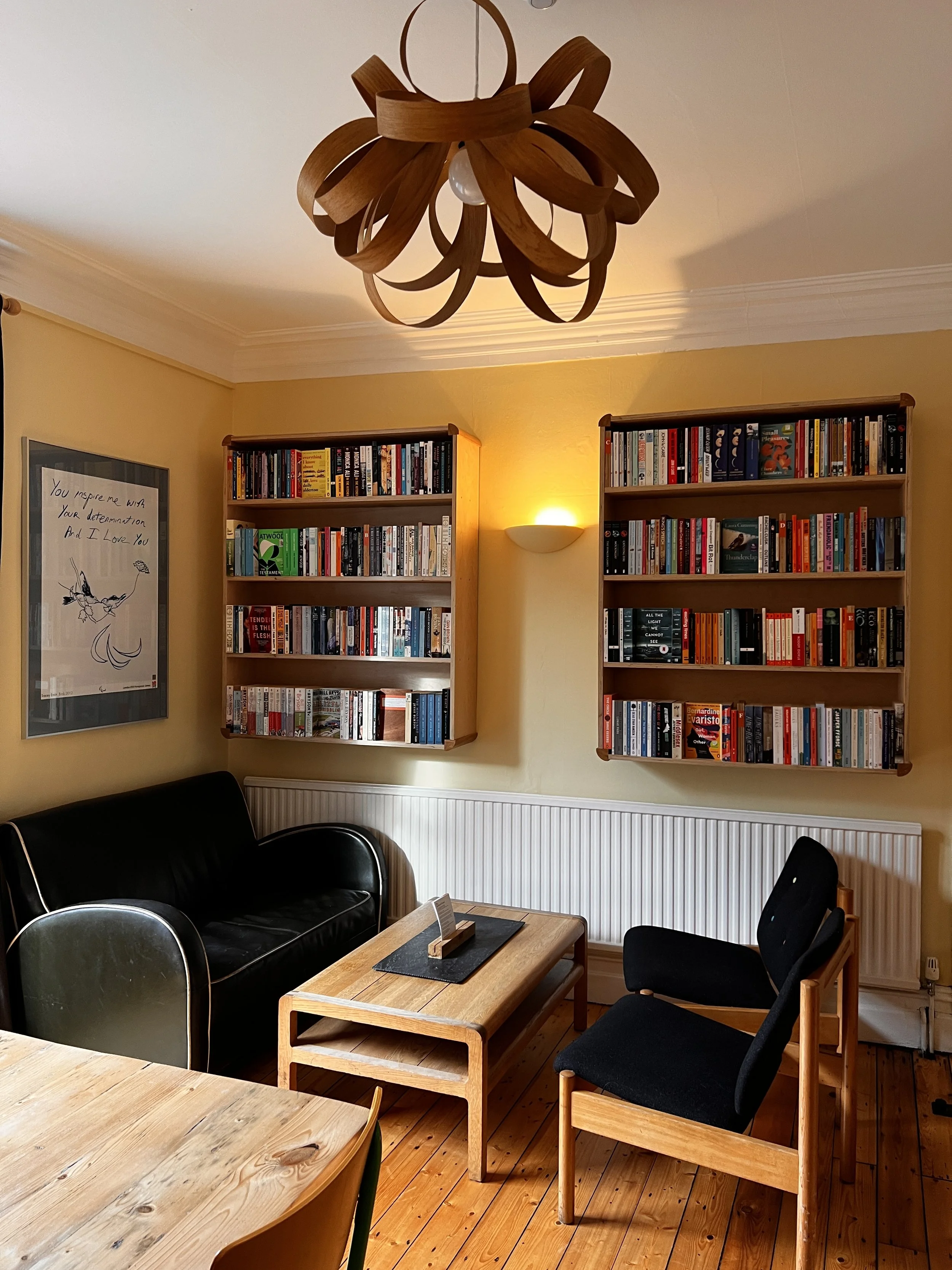 A cozy living room with a wooden coffee table, a black leather armchair, and a black upholstered chair. Two bookshelf units filled with books are mounted on a yellow wall. A framed quote and a branch illustration are on the wall. A wall-mounted light